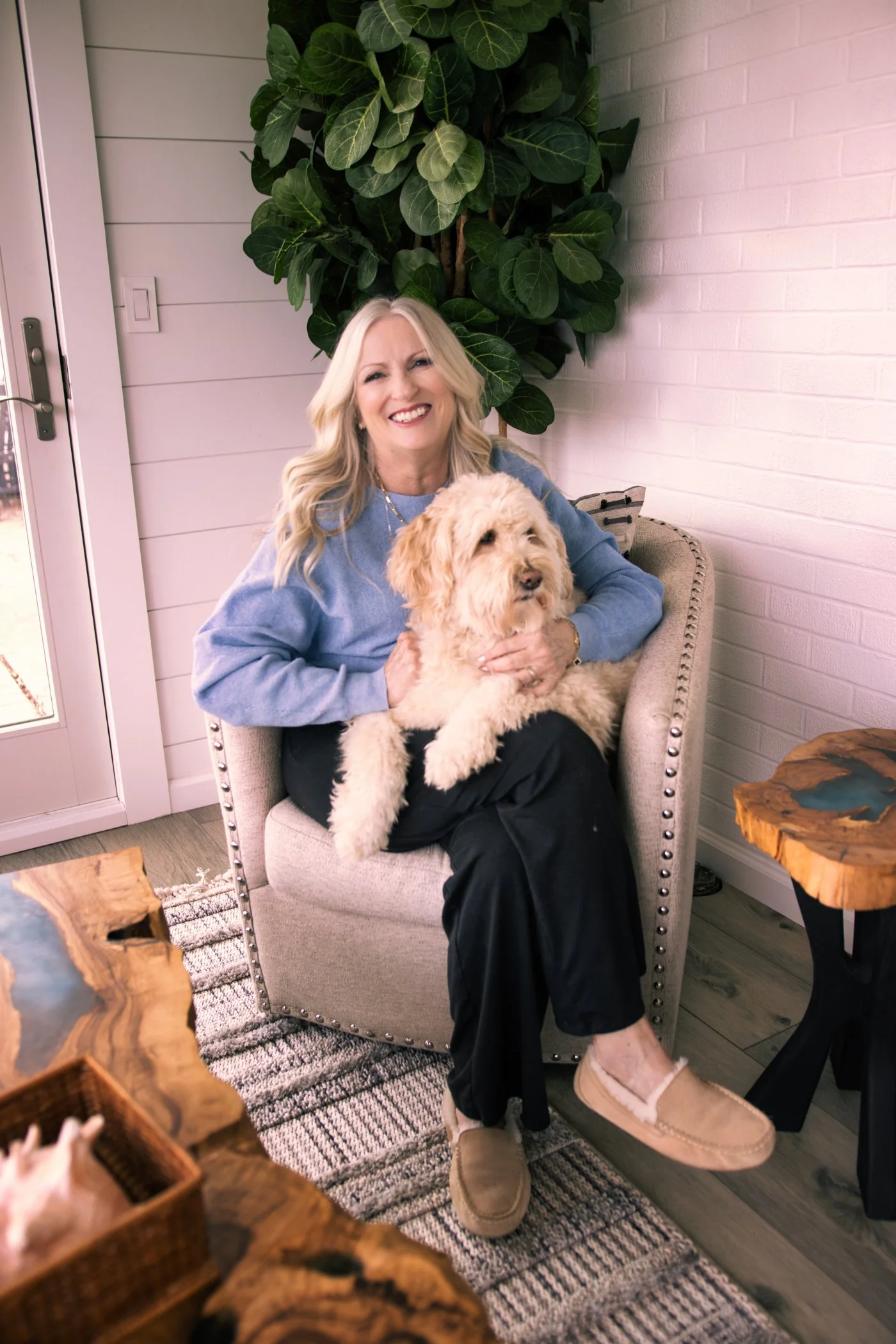 A woman with blonde hair smiling and sitting on a beige armchair, holding a fluffy cream-colored dog. She is wearing a light blue sweater, black pants, and beige slippers. There are wooden side tables near her. Behind her is a large green plant, a wh