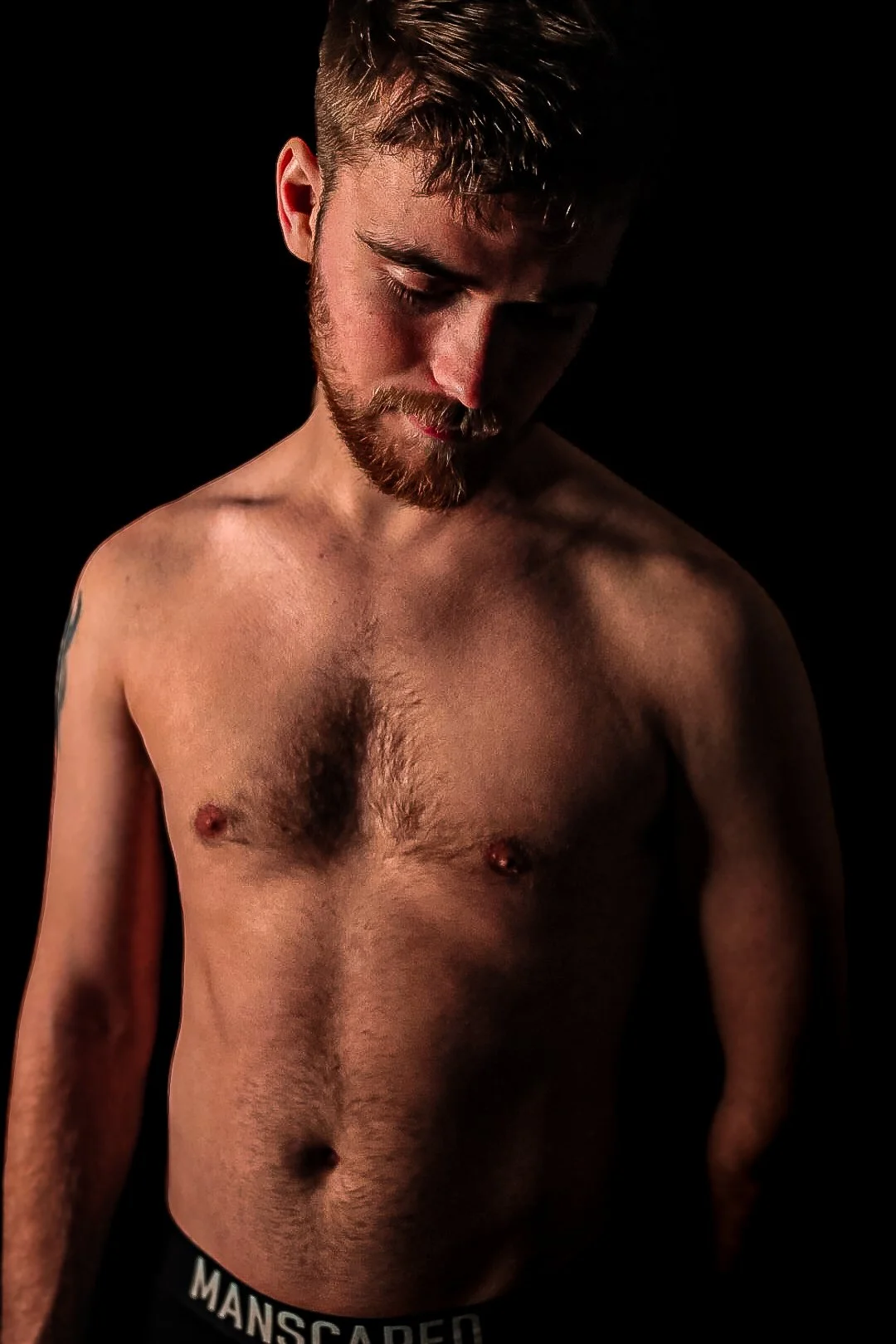 A shirtless man with a beard looking downward against a black background.