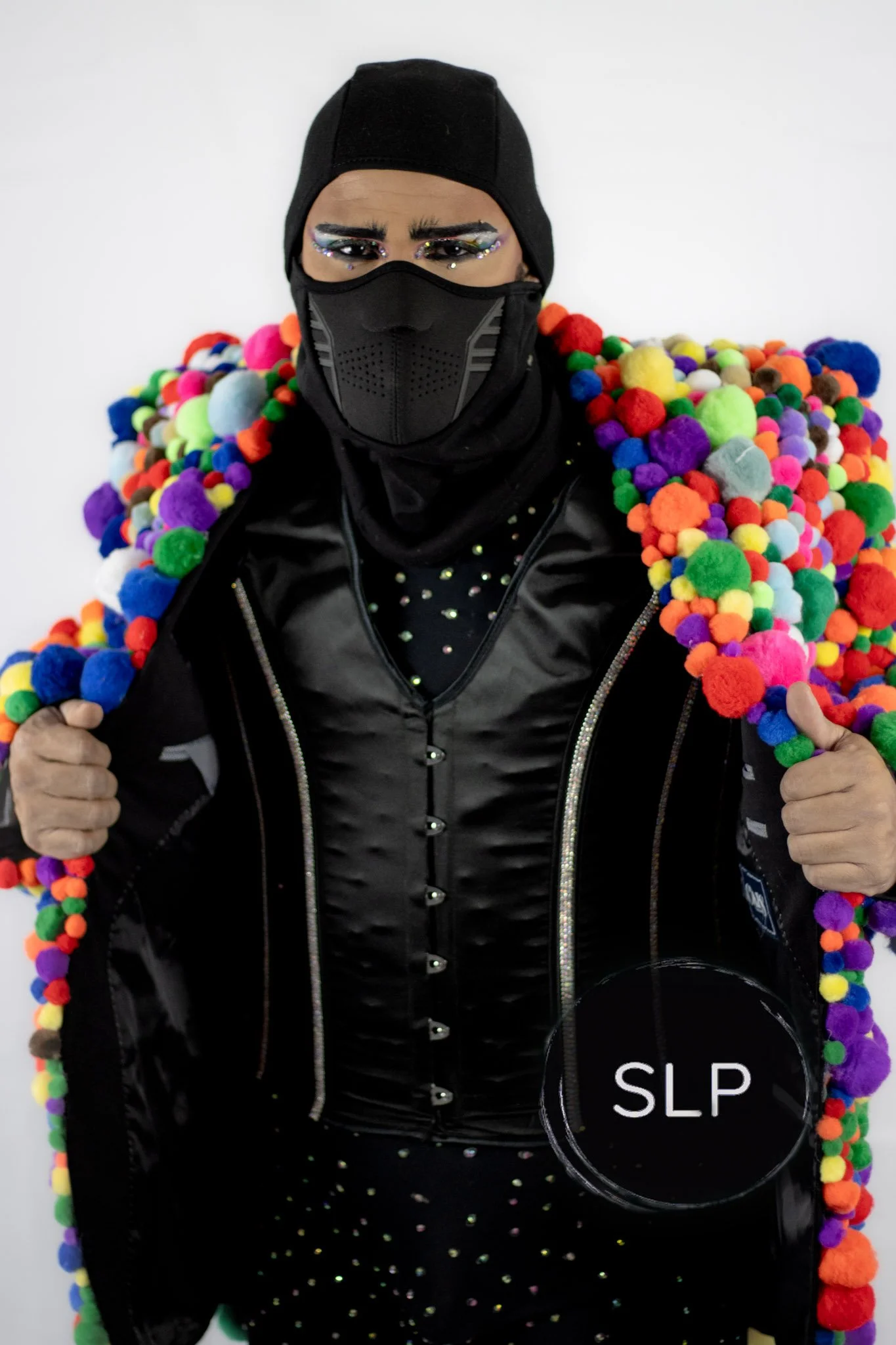Person in black winter clothing with a face mask, black beanie, and eye makeup with rhinestones, holding colorful pom-pom decorations on their shoulders and arms.