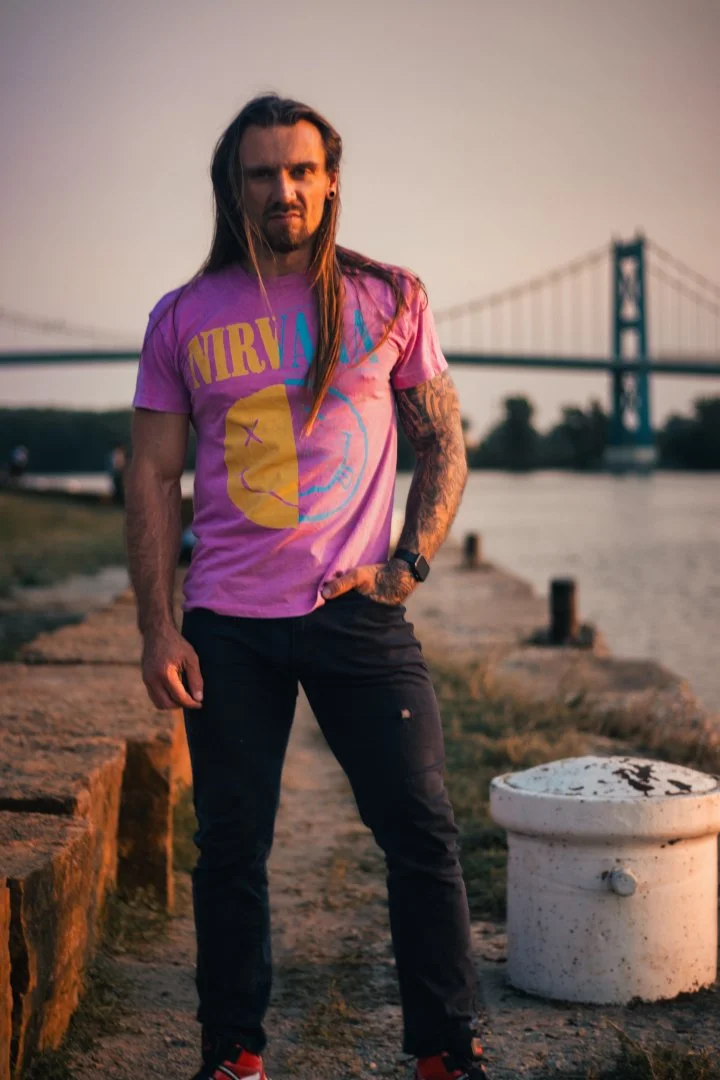 A man with long hair and tattoos wearing a pink Nirvana t-shirt and black pants standing on a riverside walkway with a bridge in the background during sunset.