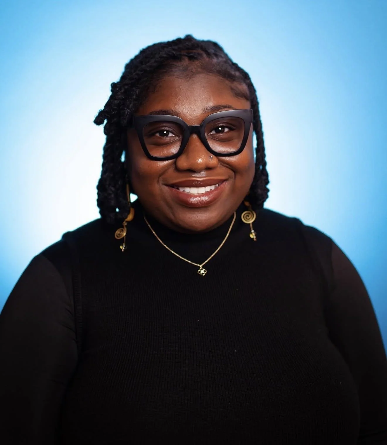 Portrait of a smiling woman wearing black glasses, gold earrings, a gold necklace, and a black top, standing against a blue gradient background.