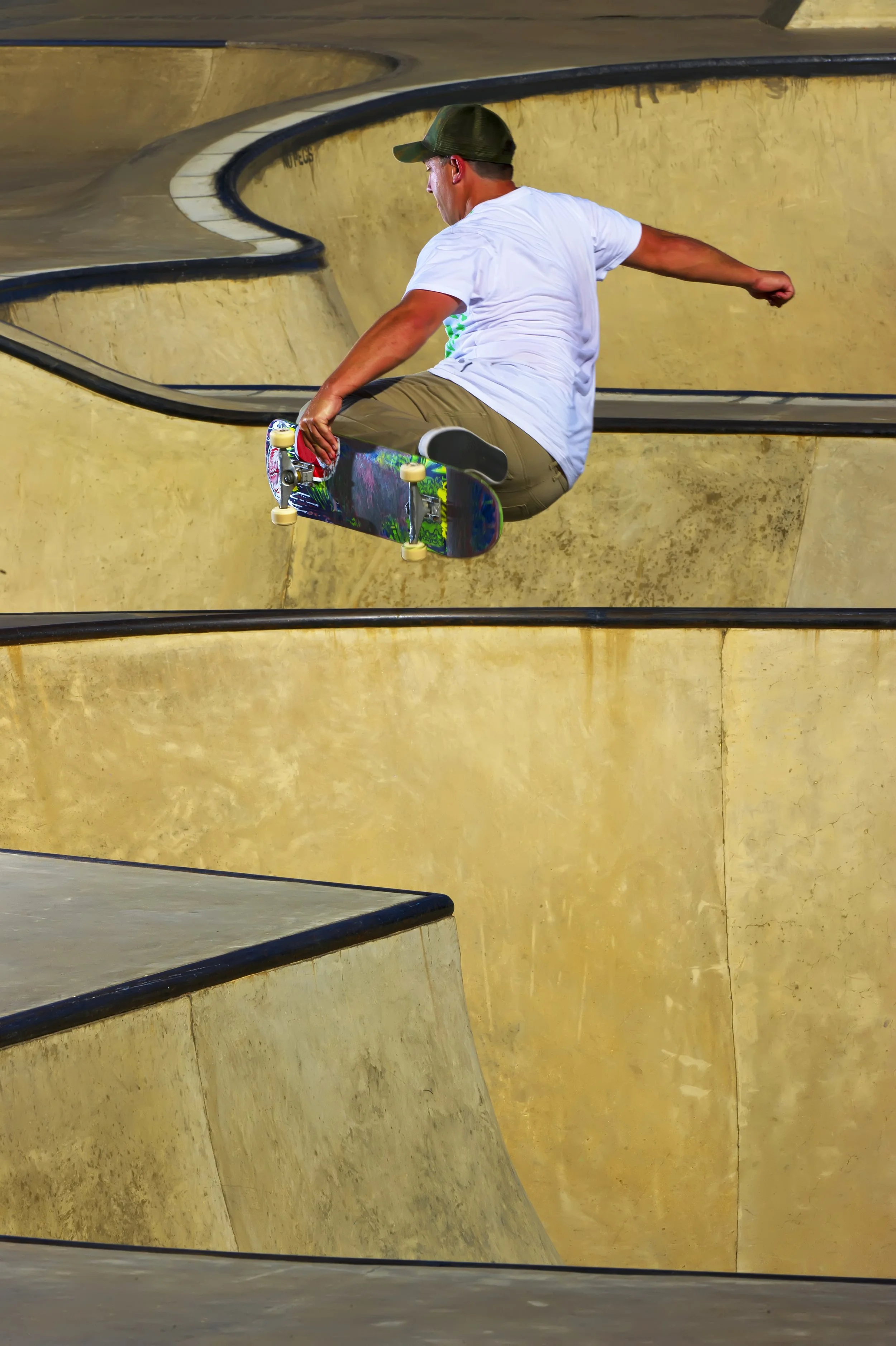 A skateboarder performing an aerial trick in a concrete skatepark bowl, wearing a white T-shirt, tan pants, and a green cap.
