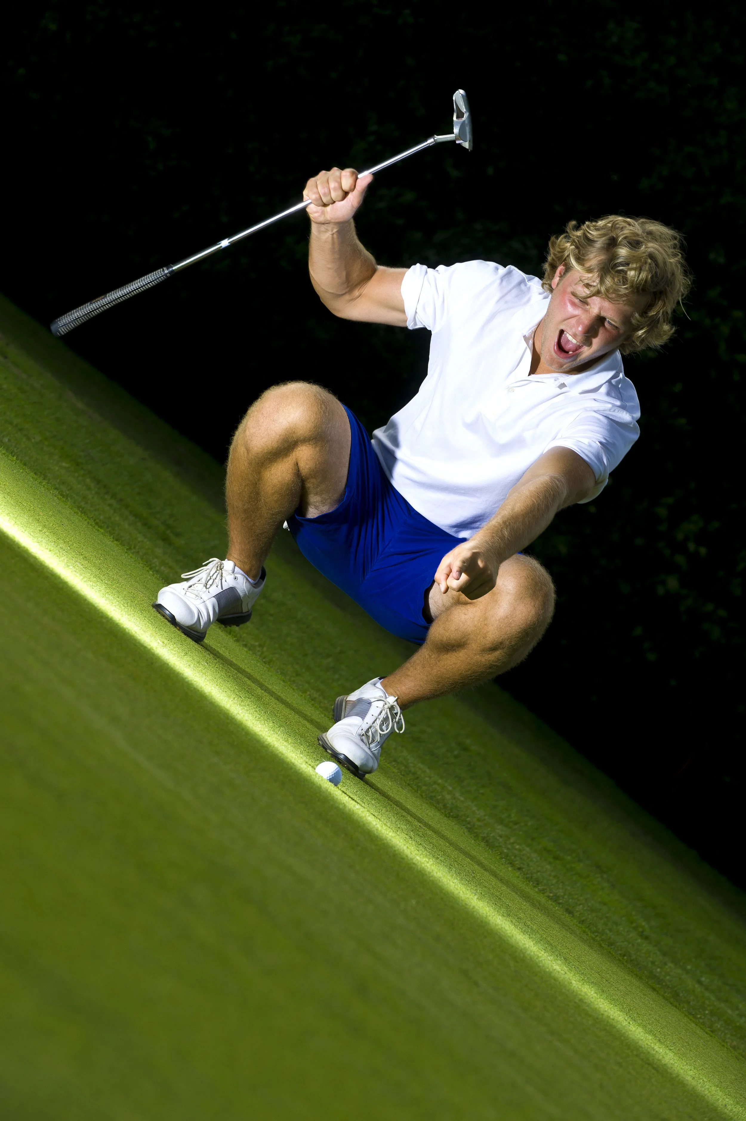 A man with curly blond hair falling backward on a golf course, holding a golf club, with a golf ball near his foot, night scene with a dark background.