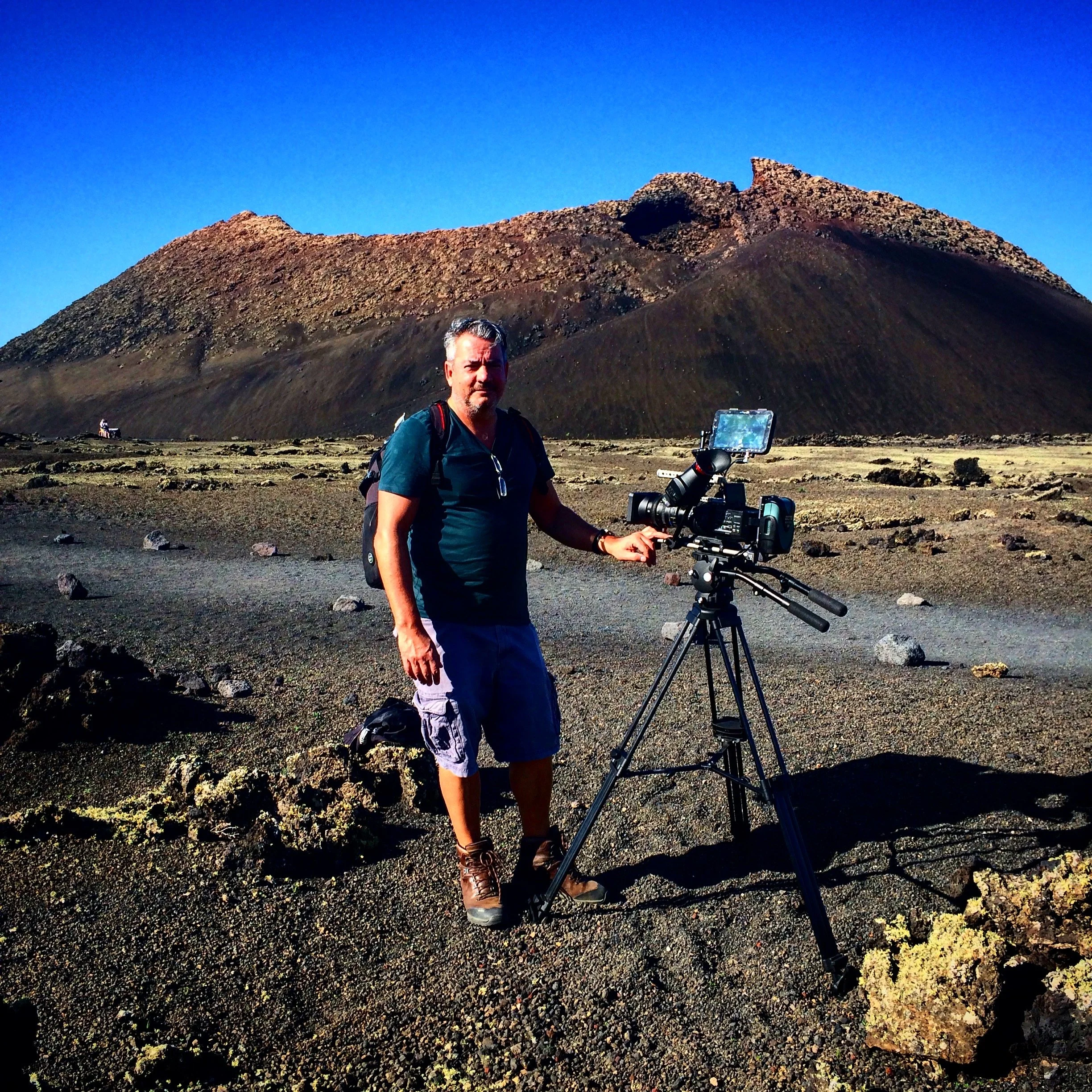 A man standing outdoors near a mountain with a bright blue sky in the background, with a camera on a tripod in front of him.