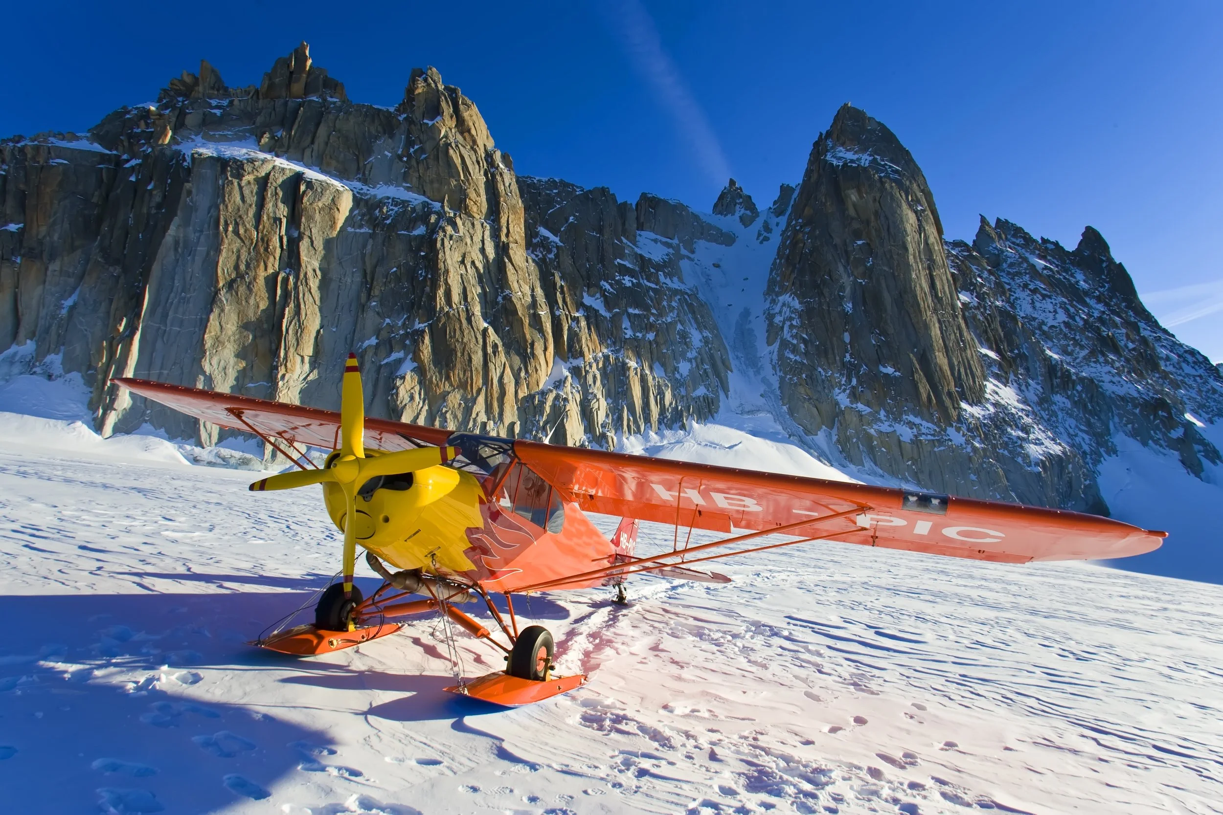 A red and yellow ultralight airplane on snowy ground with rugged snow-capped mountains in the background under a clear blue sky.