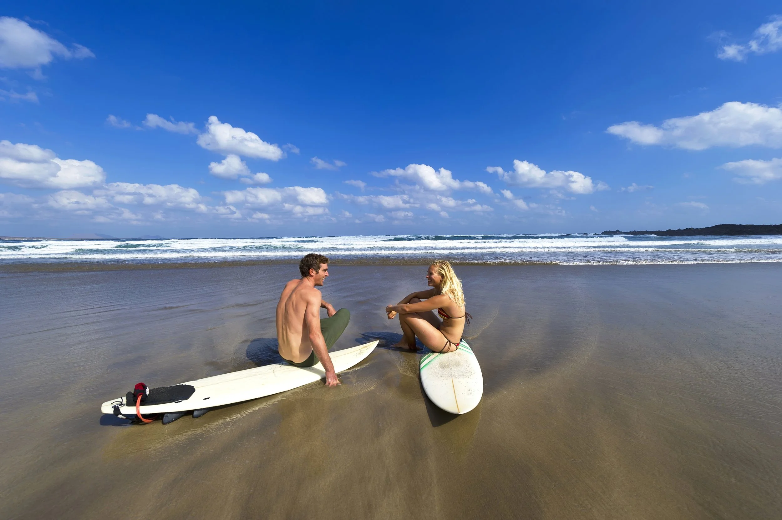 A man and woman sitting on the beach with surfboards, talking and smiling under a blue sky with clouds, with the ocean waves in the background.