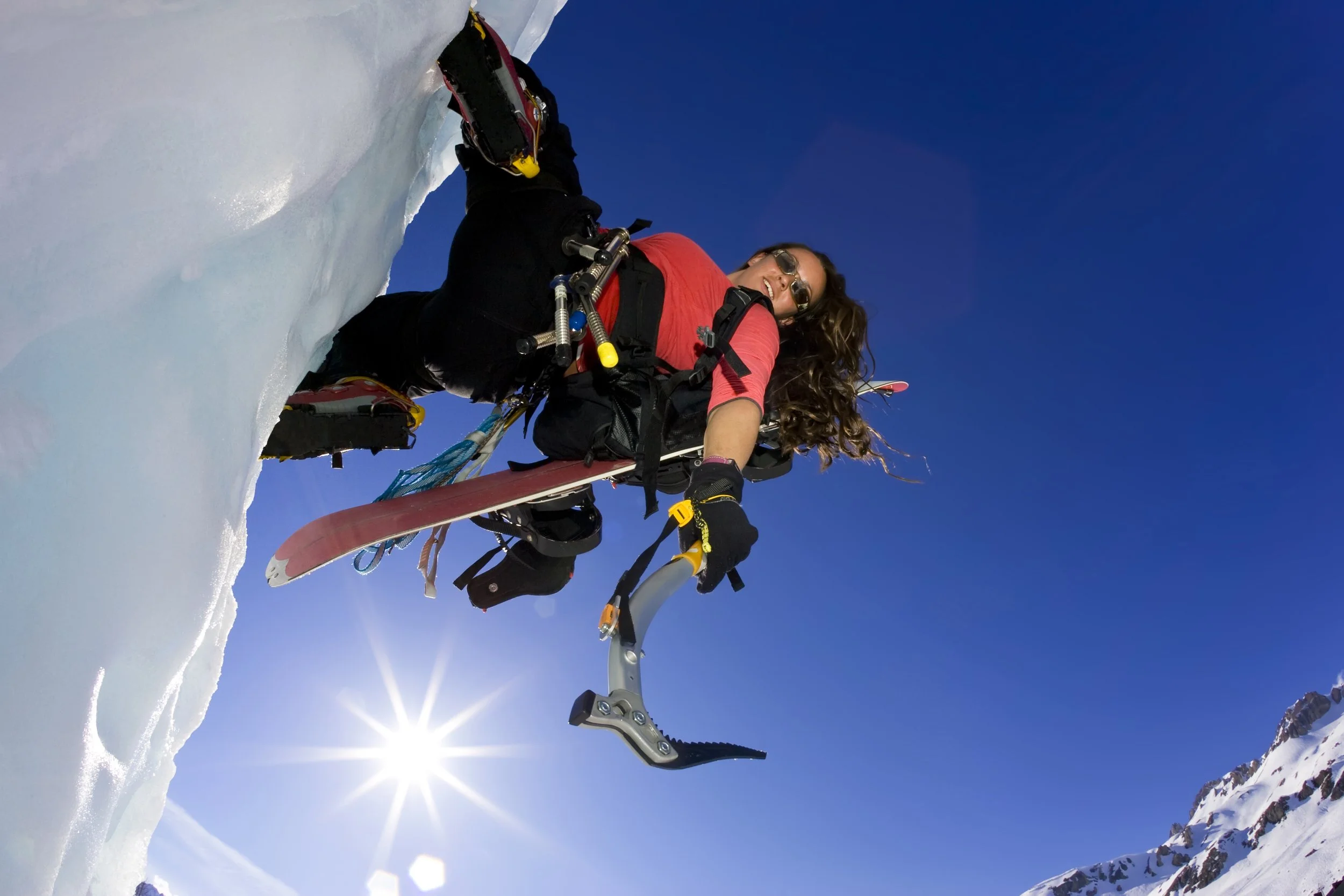 A woman lies on her stomach on a snow-covered mountain slope, wearing snow goggles and a red shirt, holding a ski pole, smiling under a bright blue sky with the sun shining and mountain peaks in the distance.
