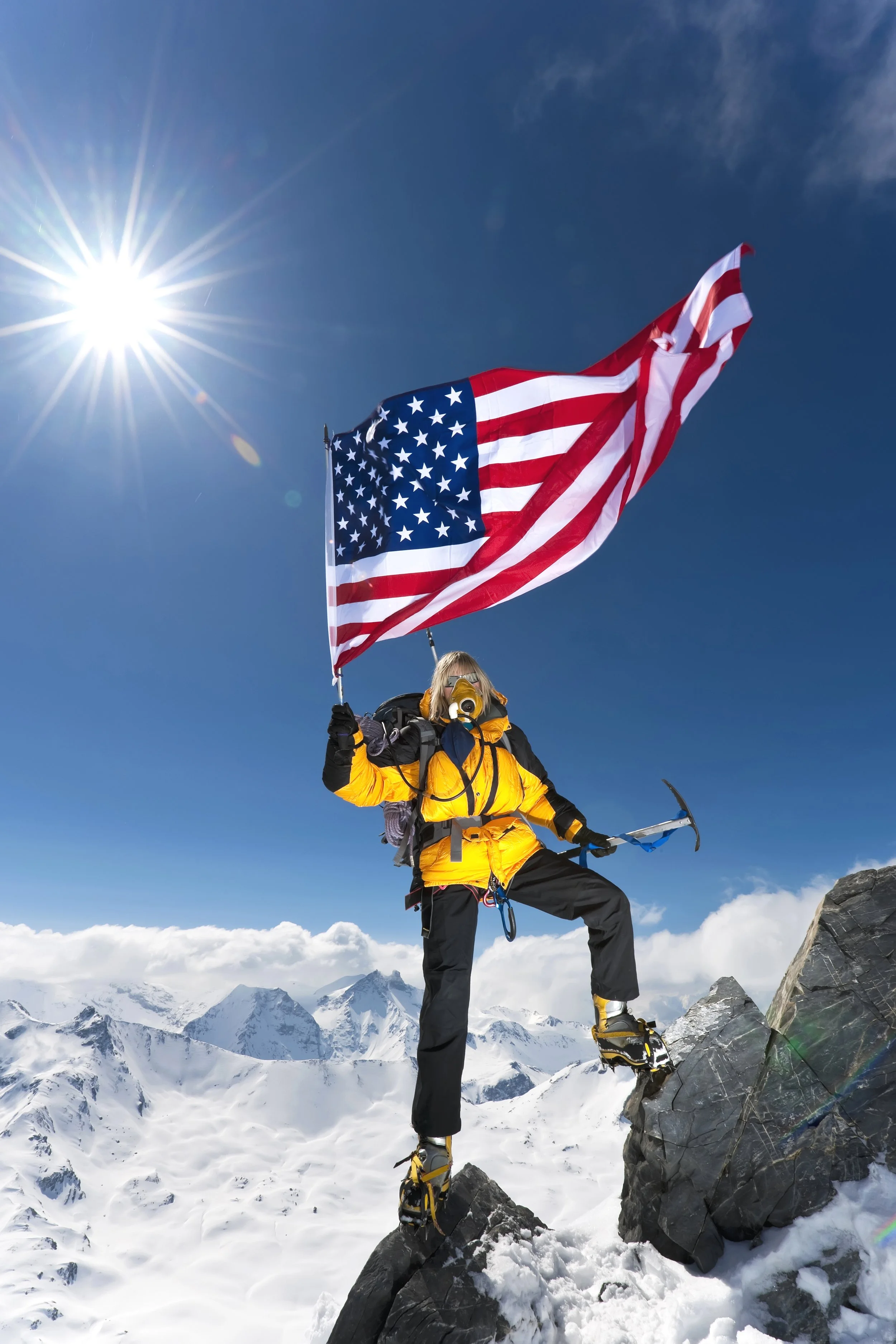 A person wearing yellow and black mountaineering gear stands on snow-covered mountain rocks, holding an American flag, with snow-capped peaks in the background and a bright sun in a clear blue sky.