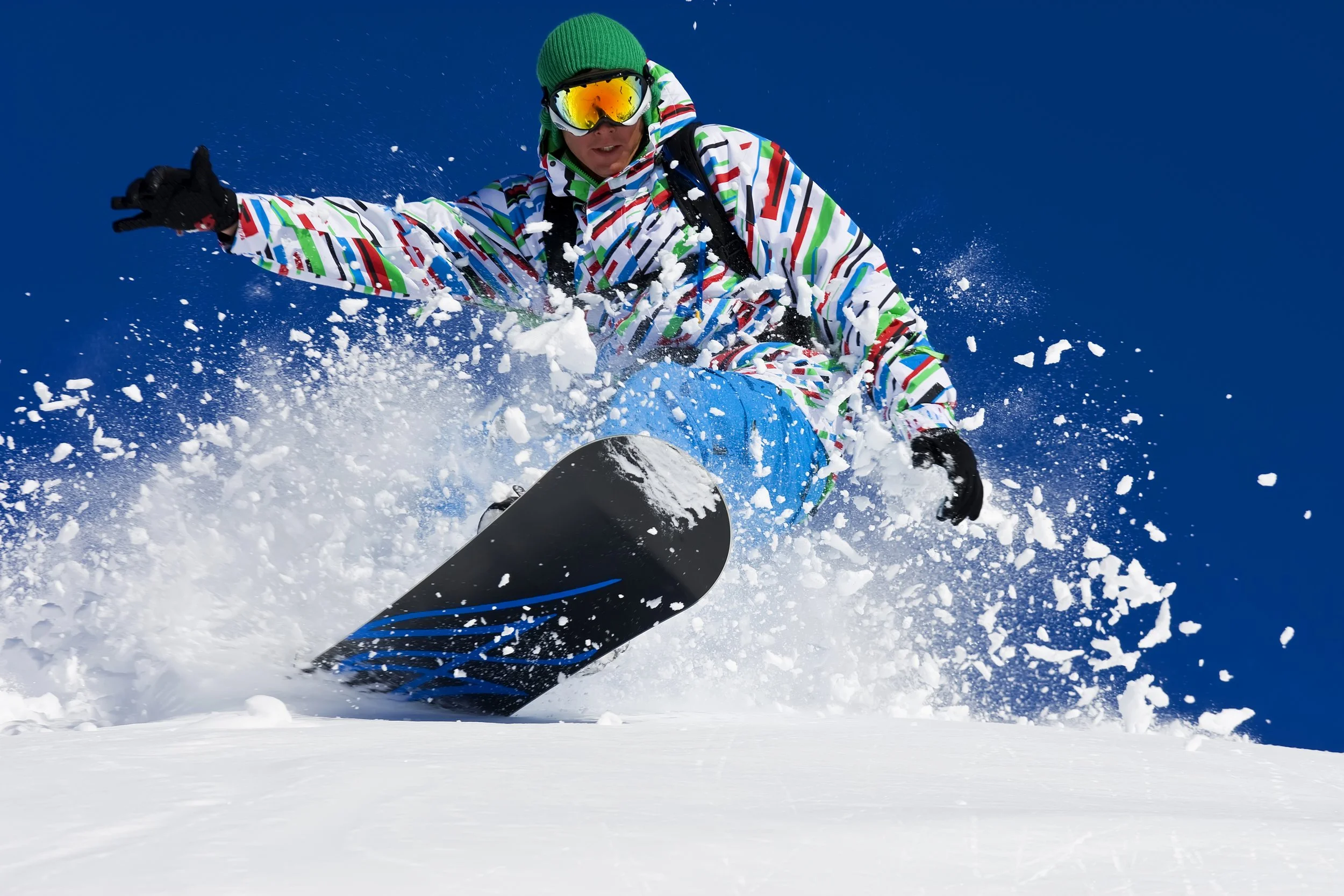 Person snowboarding down a snowy slope wearing a colorful patterned jacket, yellow goggles, green beanie, and black gloves. Snow is flying up around them against a bright blue sky.