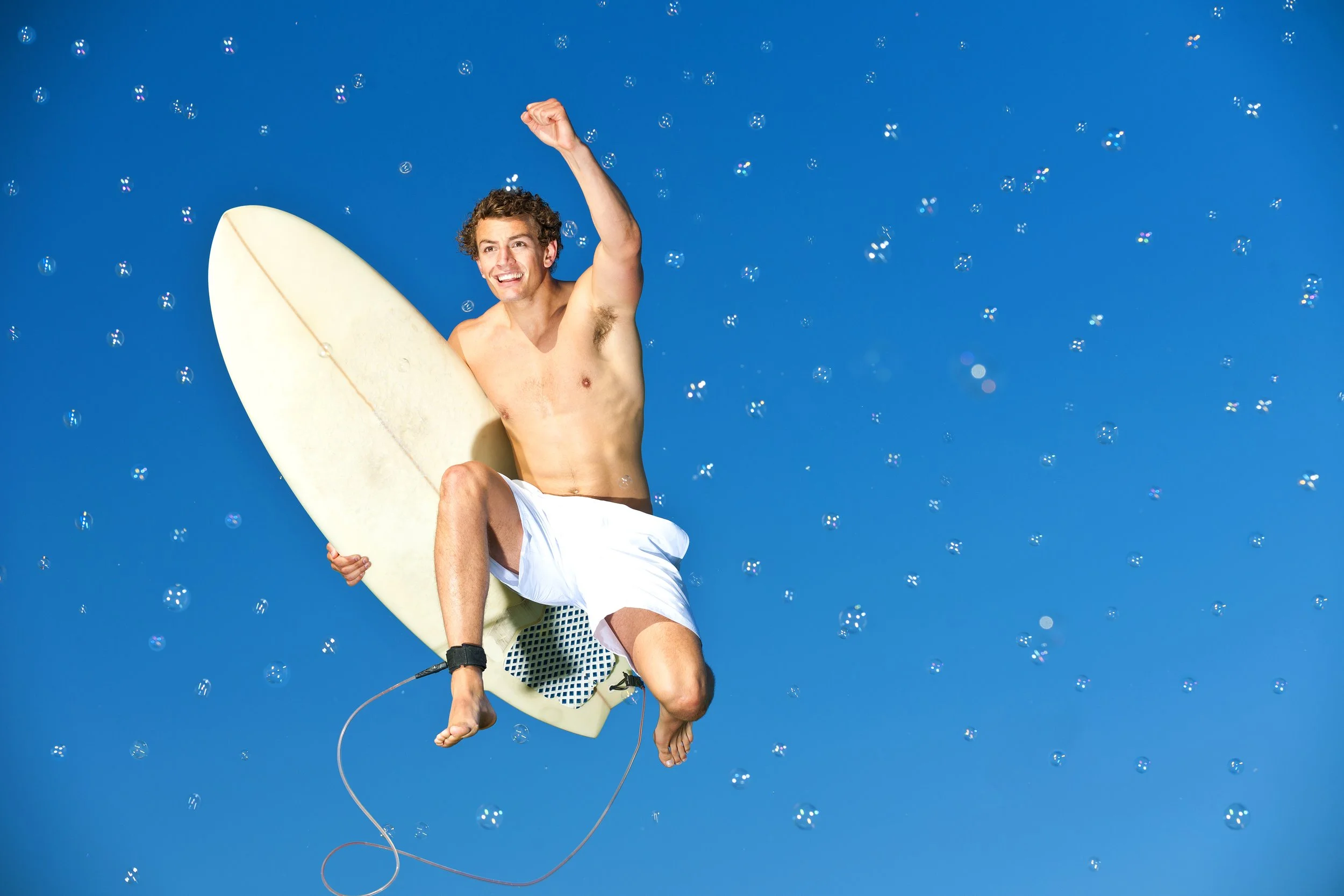 A man with curly hair, shirtless and wearing white shorts, is lying on a surfboard floating in the air above a blue sky with small floating bubbles, giving an impression of flying or floating.