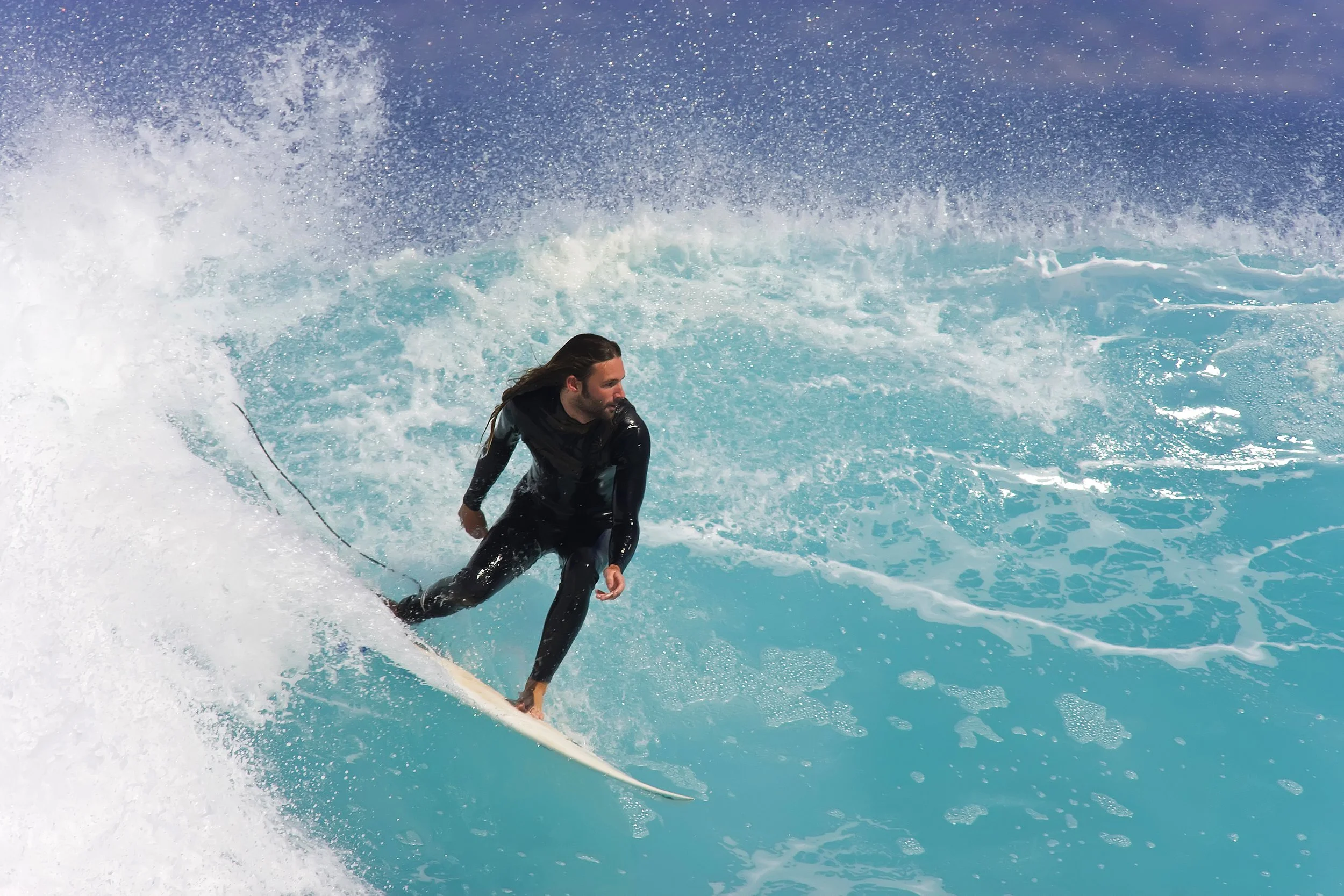 A person with long hair wakeboarding on bright blue water, holding a rope, and leaning to one side with water splashing around.