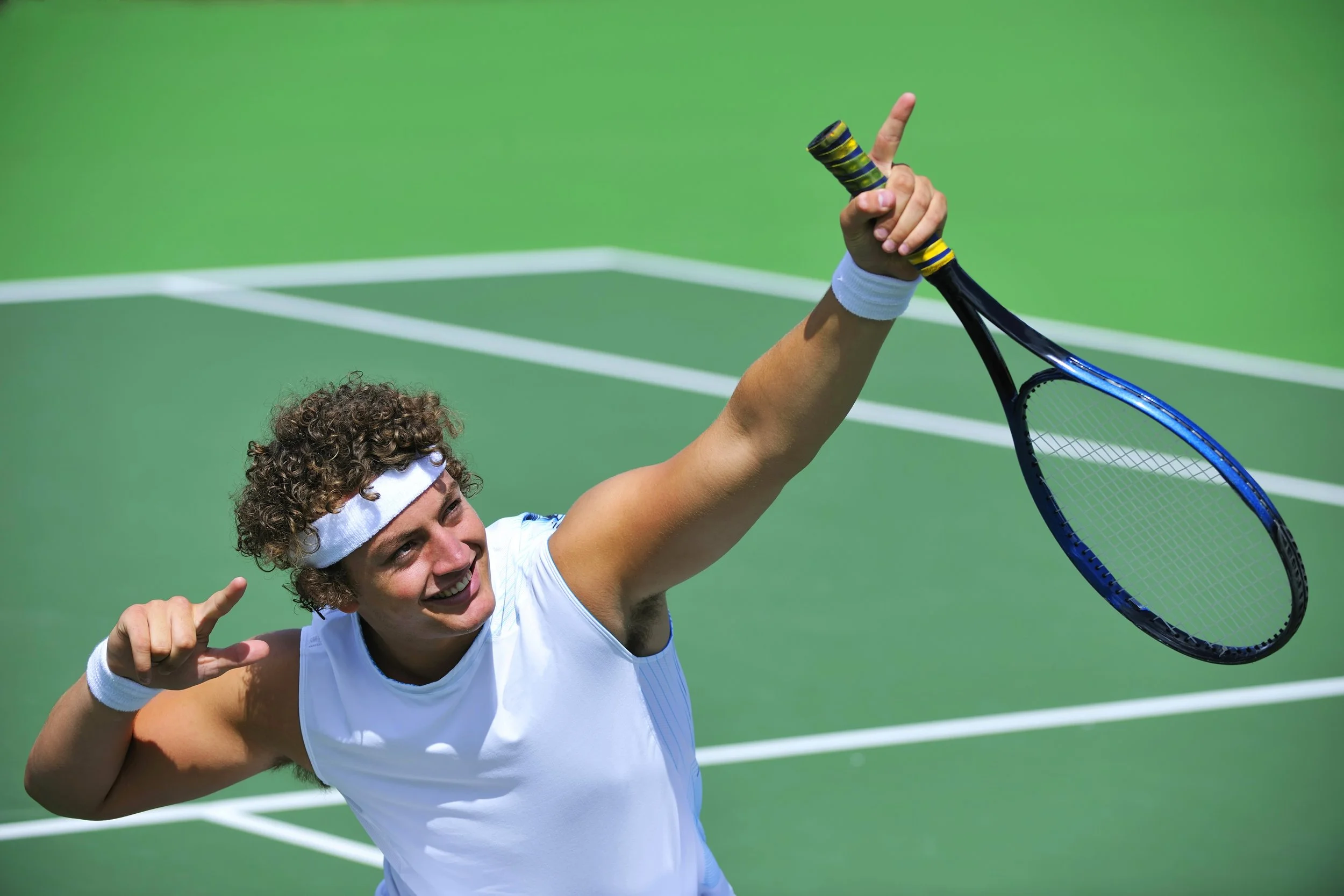 A young male tennis player celebrating on a green tennis court, holding his tennis racket up with one hand and pointing with his index finger. He is wearing a white sports shirt and a white headband, smiling.