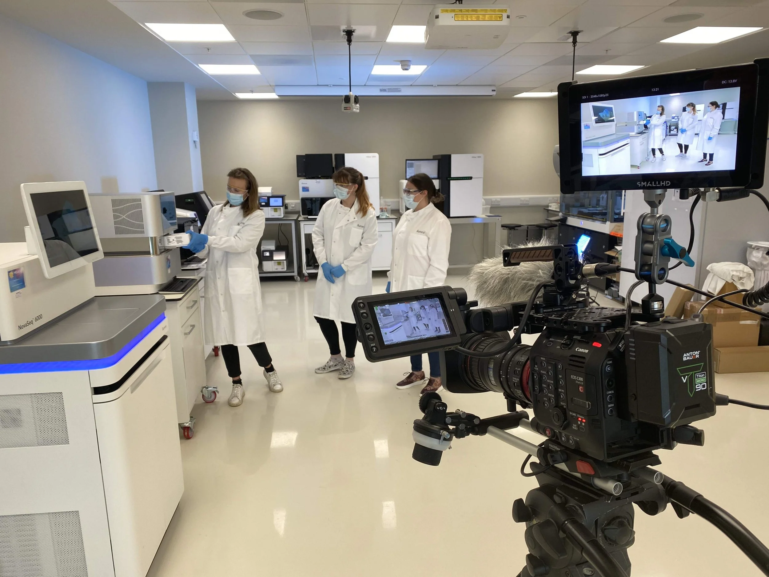 A group of four scientists or lab technicians wearing white lab coats, face masks, and gloves in a medical or research laboratory. They are standing near lab equipment and appear to be preparing or analyzing samples. A professional video camera on a tripod is filming the scene, with a monitor displaying the recording.