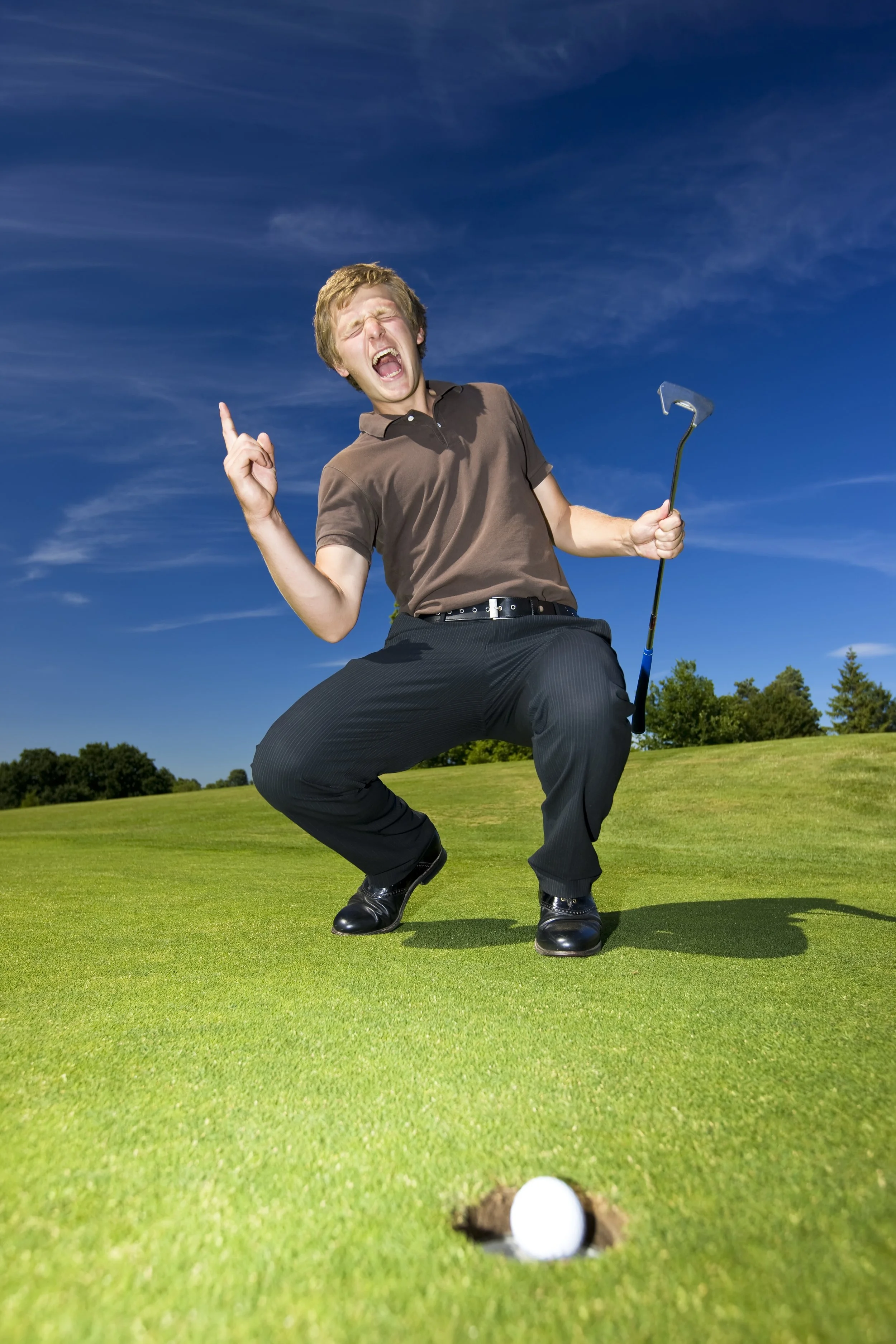 Man dressed in formal pants and button-up shirt, crouching on a golf course, holding a golf club, with a golf ball near the hole, under a clear blue sky.