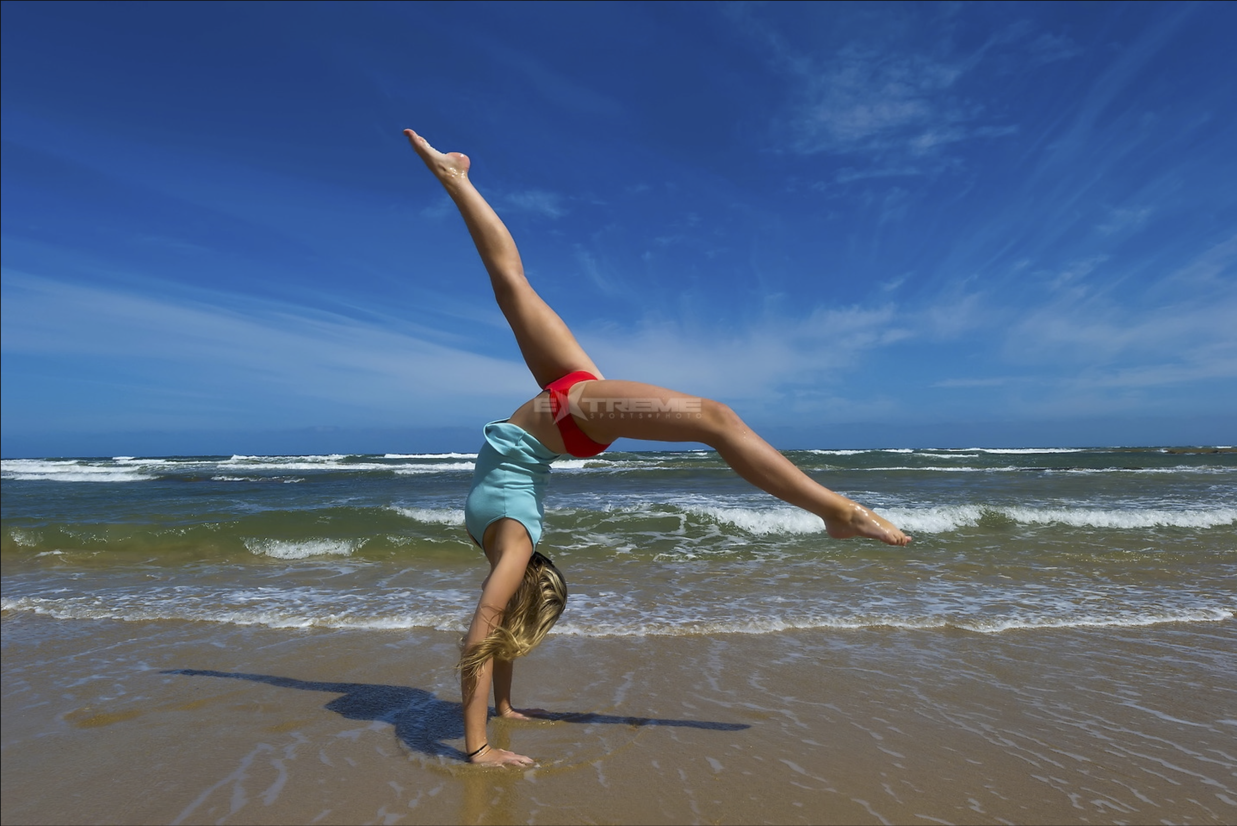 A person doing a handstand on the beach with ocean waves in the background and a clear blue sky.