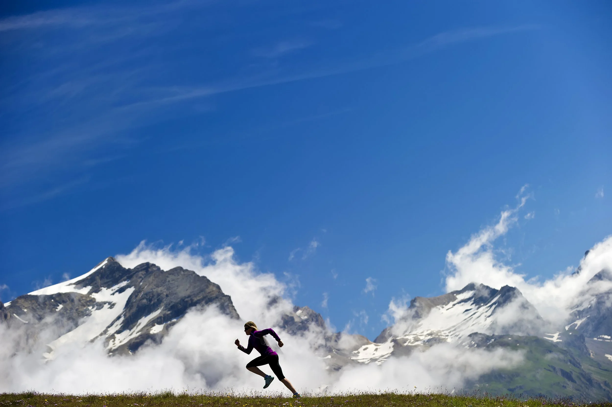 A person wearing a purple jacket and black pants running on grass in front of snow-capped mountains under a blue sky with wispy clouds.
