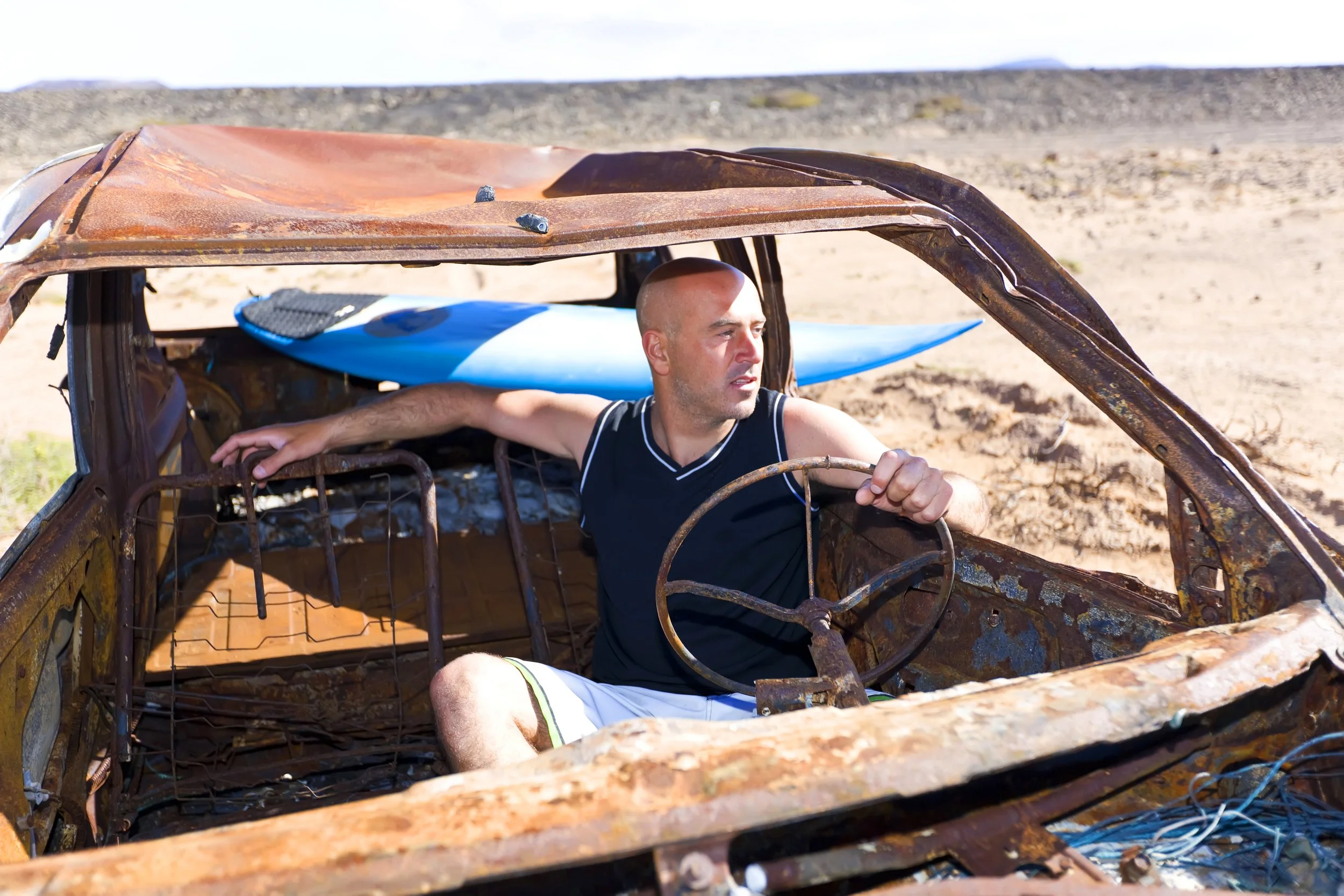 A man sitting inside a rusted, abandoned car with a blue surfboard in the background on a desert landscape.