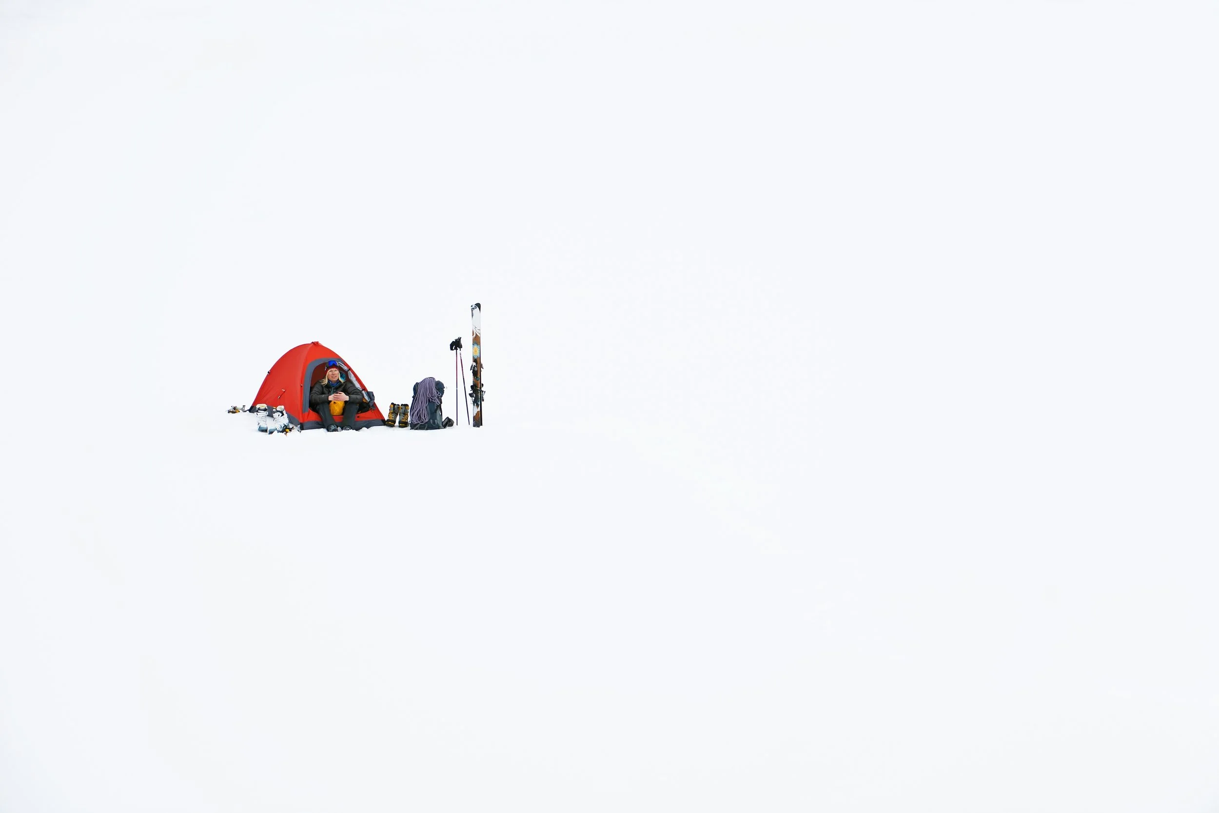 A person sitting in a small red tent in a snowy landscape, with ski equipment including skis and poles nearby.
