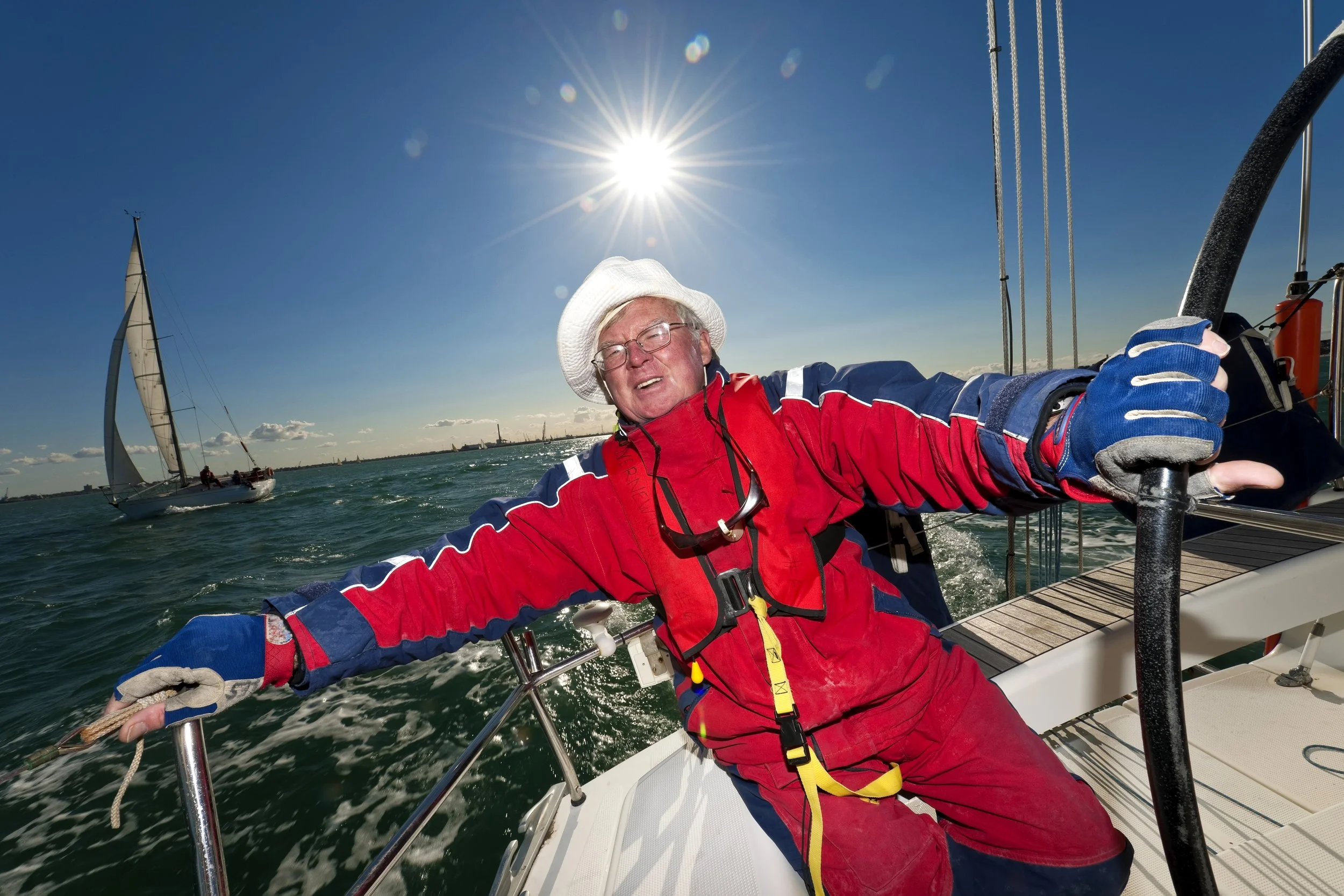 A man wearing a red and blue waterproof jacket, gloves, and a white hat is sailing on a boat. He is holding onto a railing with a bright sun overhead, and another sailboat is visible in the distance on the water.