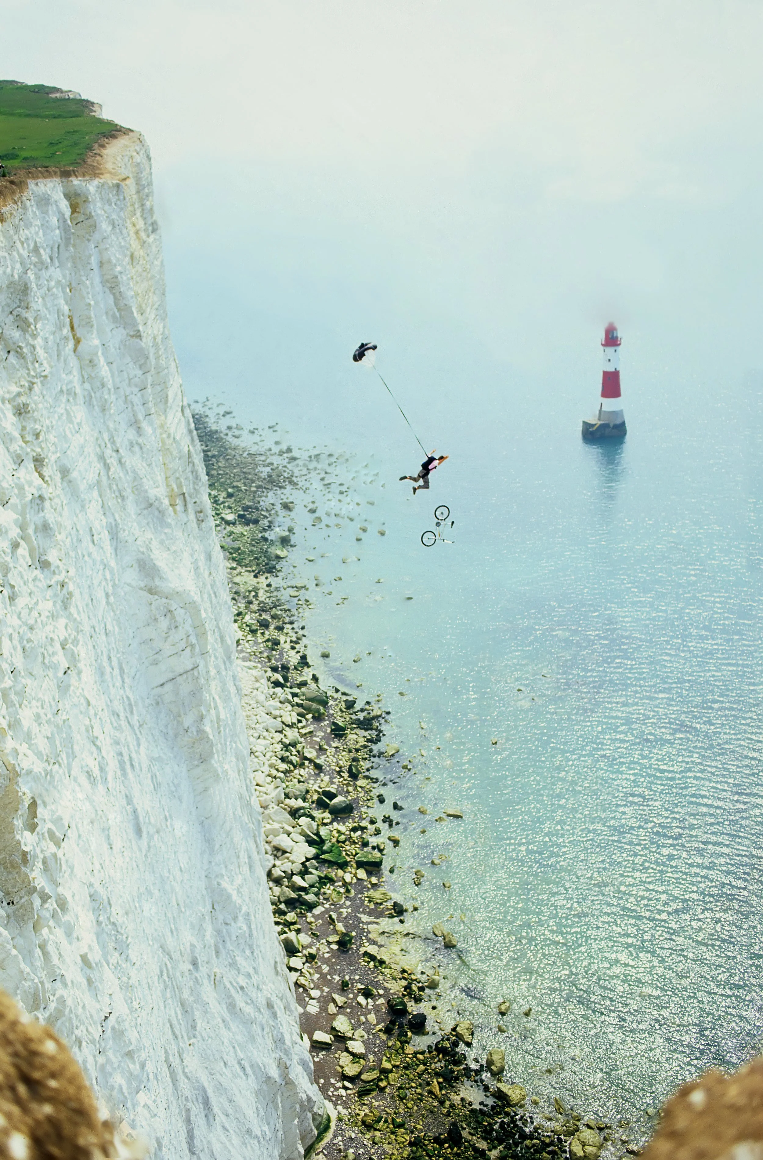 A person hanging from a rope over white chalk cliffs near the ocean, with a person riding a bicycle and a lighthouse in the background.