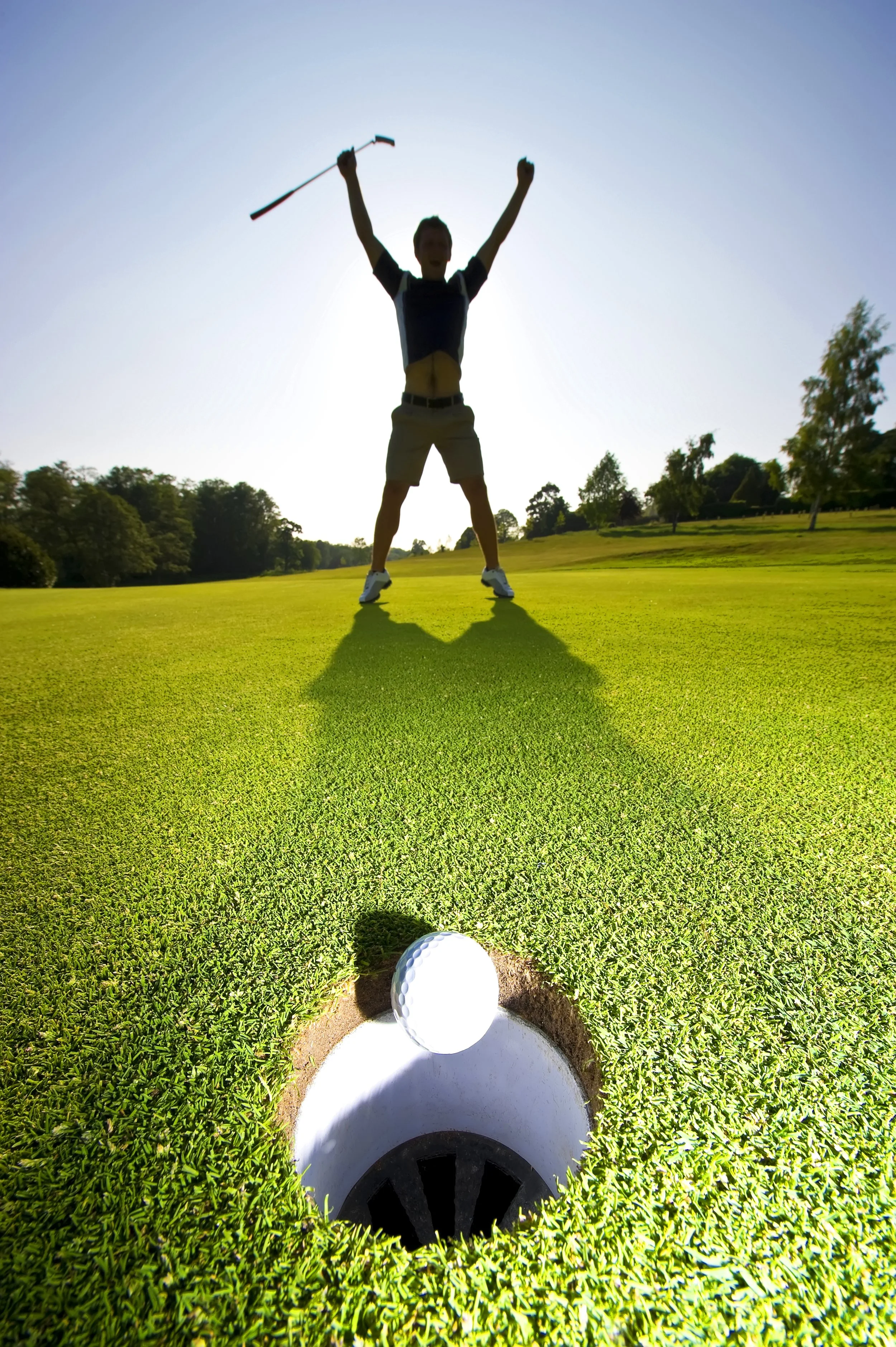 A person celebrating with their arms raised on a golf course, viewed from behind a golf ball in a sand trap, with a golf hole and flag in the background.