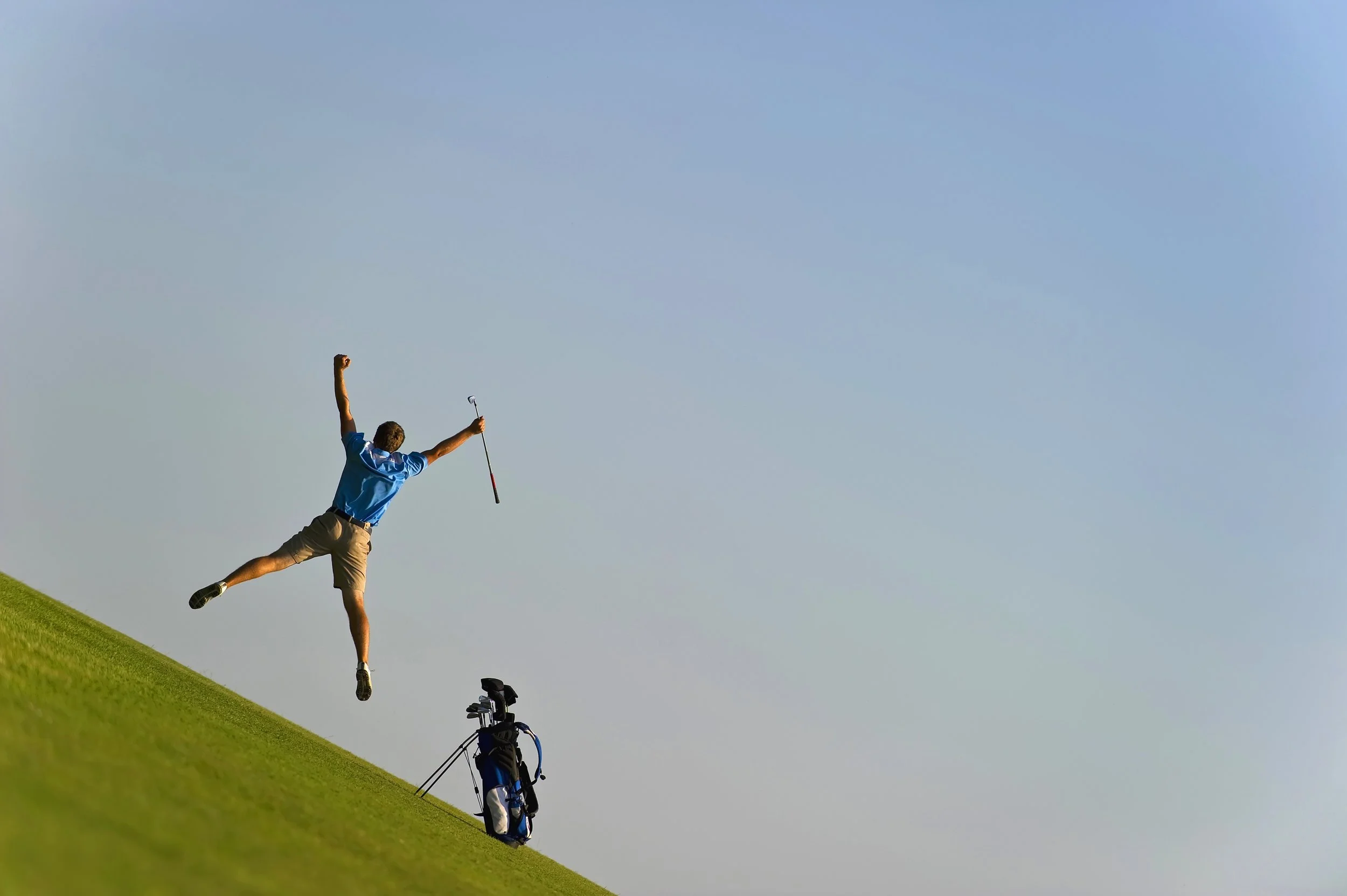 A person in a blue shirt and beige shorts is jumping in the air on a golf course, holding a golf club in one hand, with a golf bag and golf clubs on the ground nearby. The sky is clear and blue.