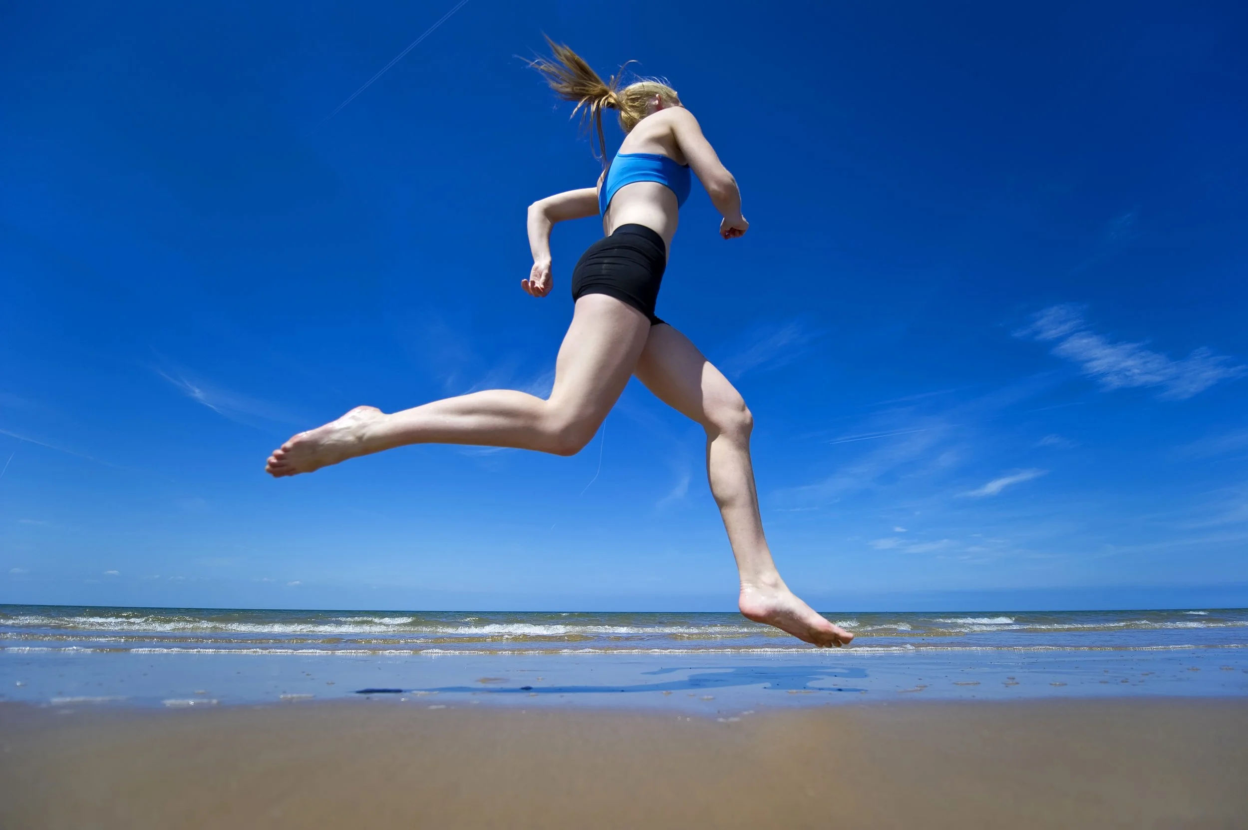 A woman running on the beach under a clear blue sky, wearing a blue sports bra and black shorts.