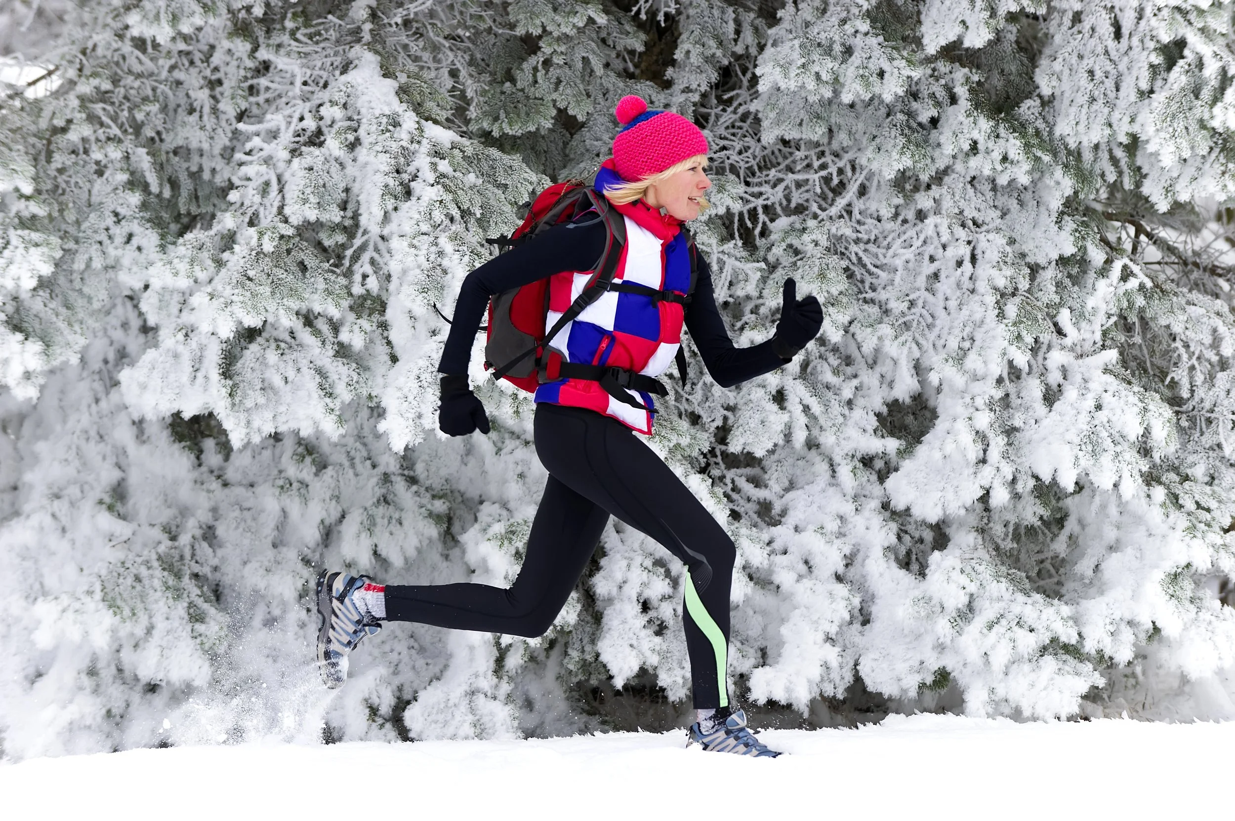 A woman running through a snowy forest wearing a bright pink knit hat with a pom-pom, a red, white, and blue jacket, black leggings, and grey running shoes with blue accents. She has a red backpack and is giving a thumbs-up along with a smile.