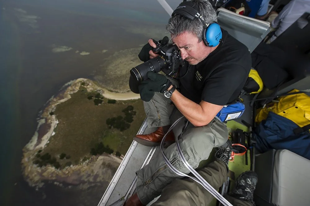 A man sitting on the edge of an aircraft, taking a photo of a small island below, with headphones on and camera in hand.