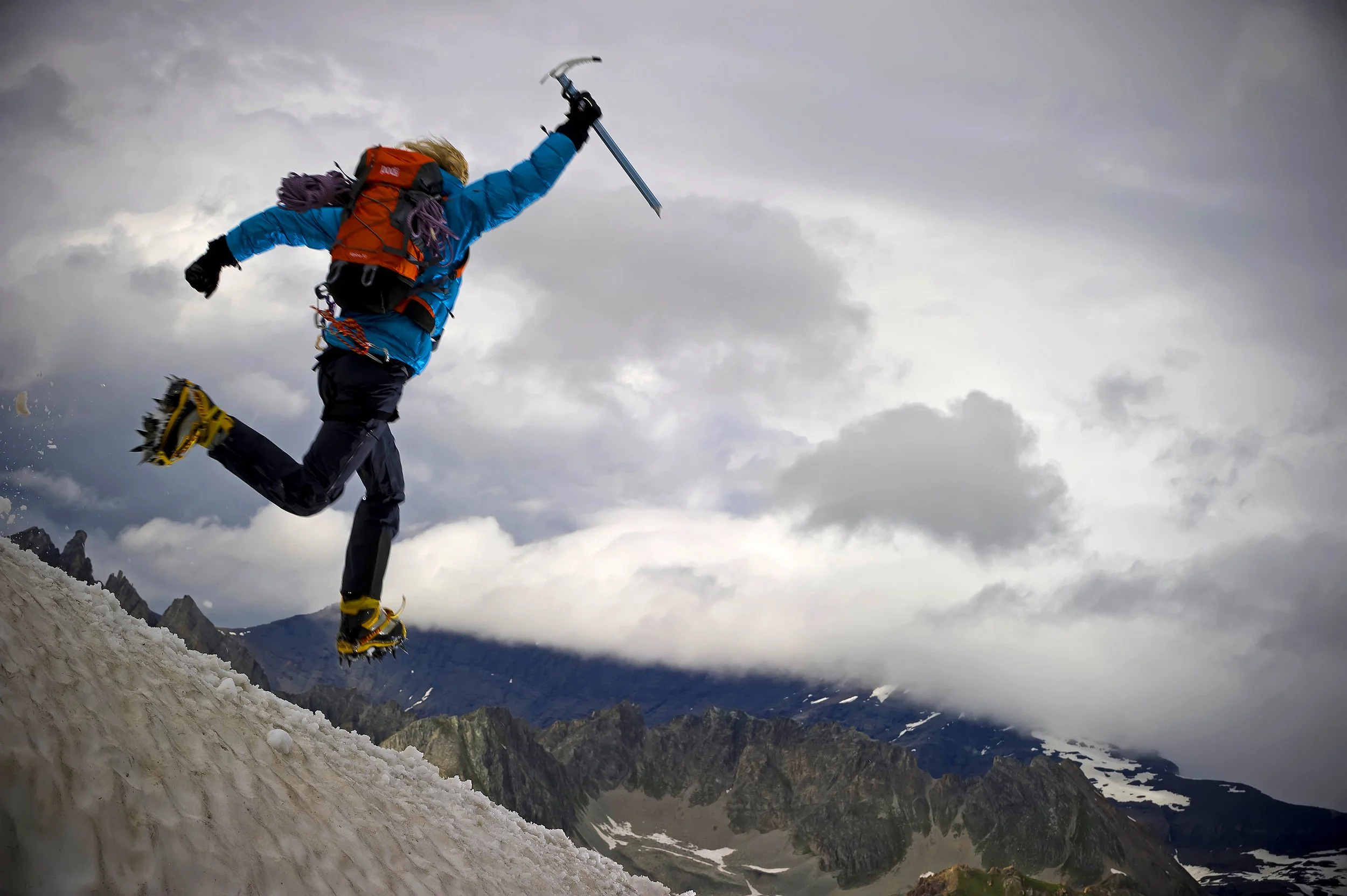 A person wearing a blue jacket, black pants, and yellow snowshoes is snowshoeing on a snowy mountain trail, with mountains and cloudy sky in the background.