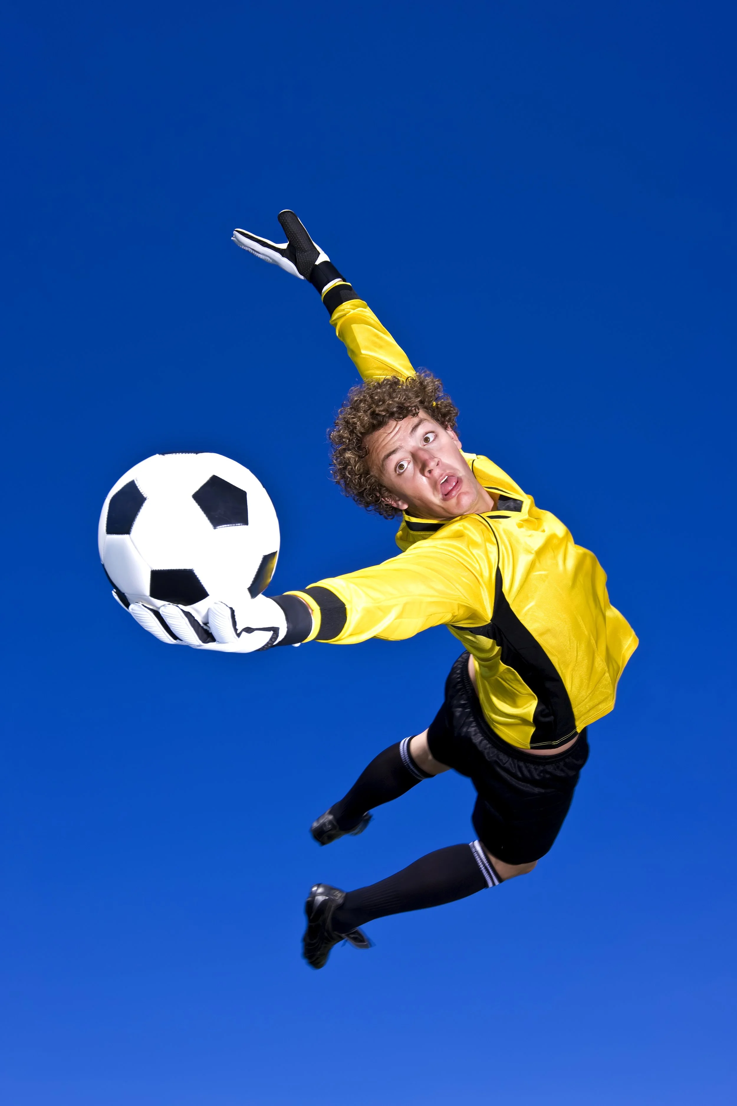 A soccer goalkeeper in a yellow jersey and shorts is diving in the air to catch a black and white soccer ball against a bright blue background.
