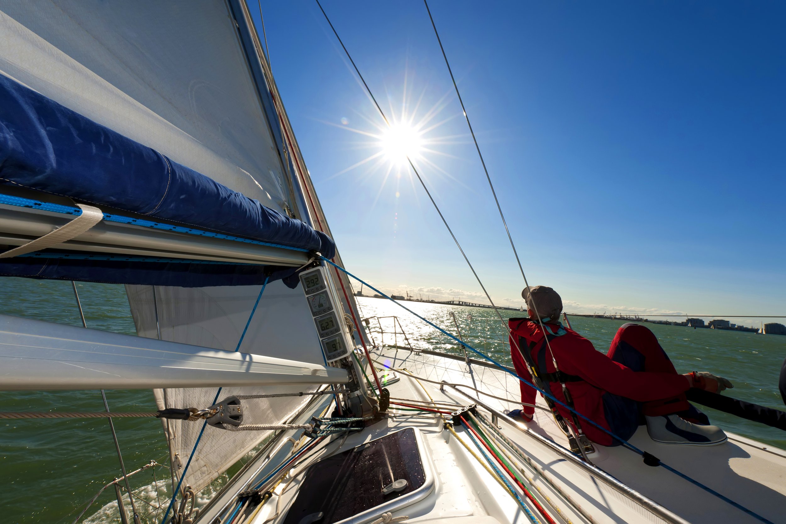 A sailboat on the water with the sun shining brightly overhead, a crew member in a red jacket leaning back on the boat, and the city skyline visible in the distance.