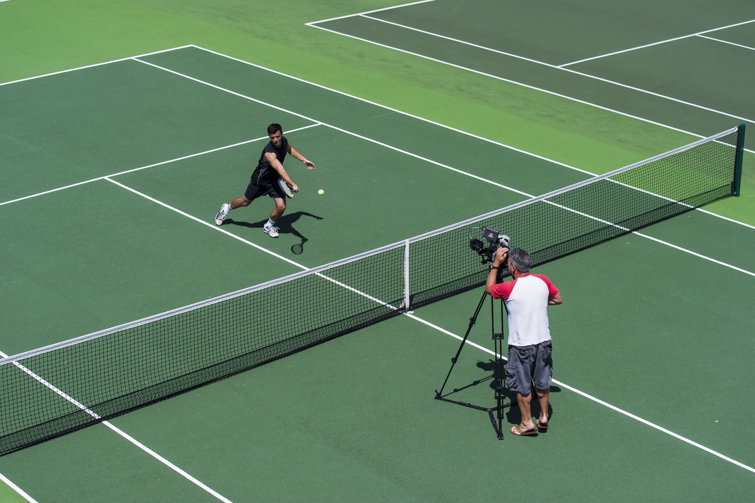 A man playing tennis on an outdoor court, preparing to hit a tennis ball near the net, with a photographer filming from behind.