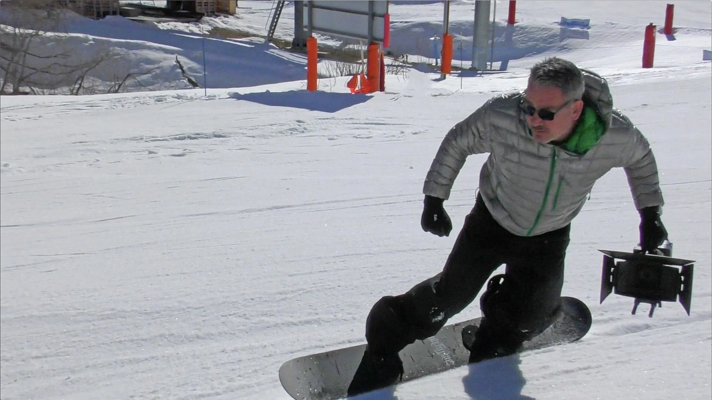 A man in sunglasses and winter clothing is snowboarding on a snowy slope, with a small black sled nearby.