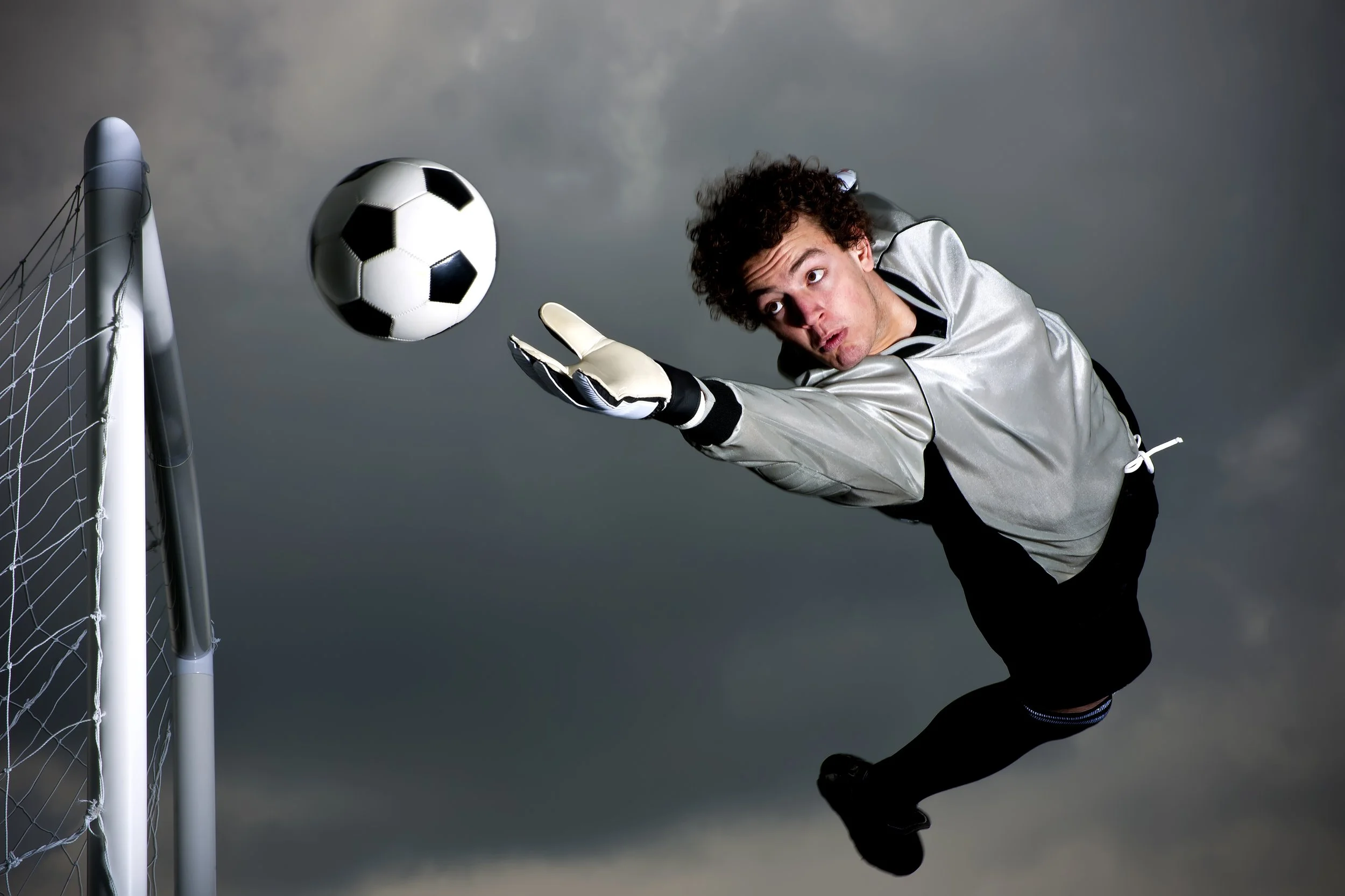 A soccer goalkeeper diving to save a black and white soccer ball near the goalpost on a cloudy sky background.