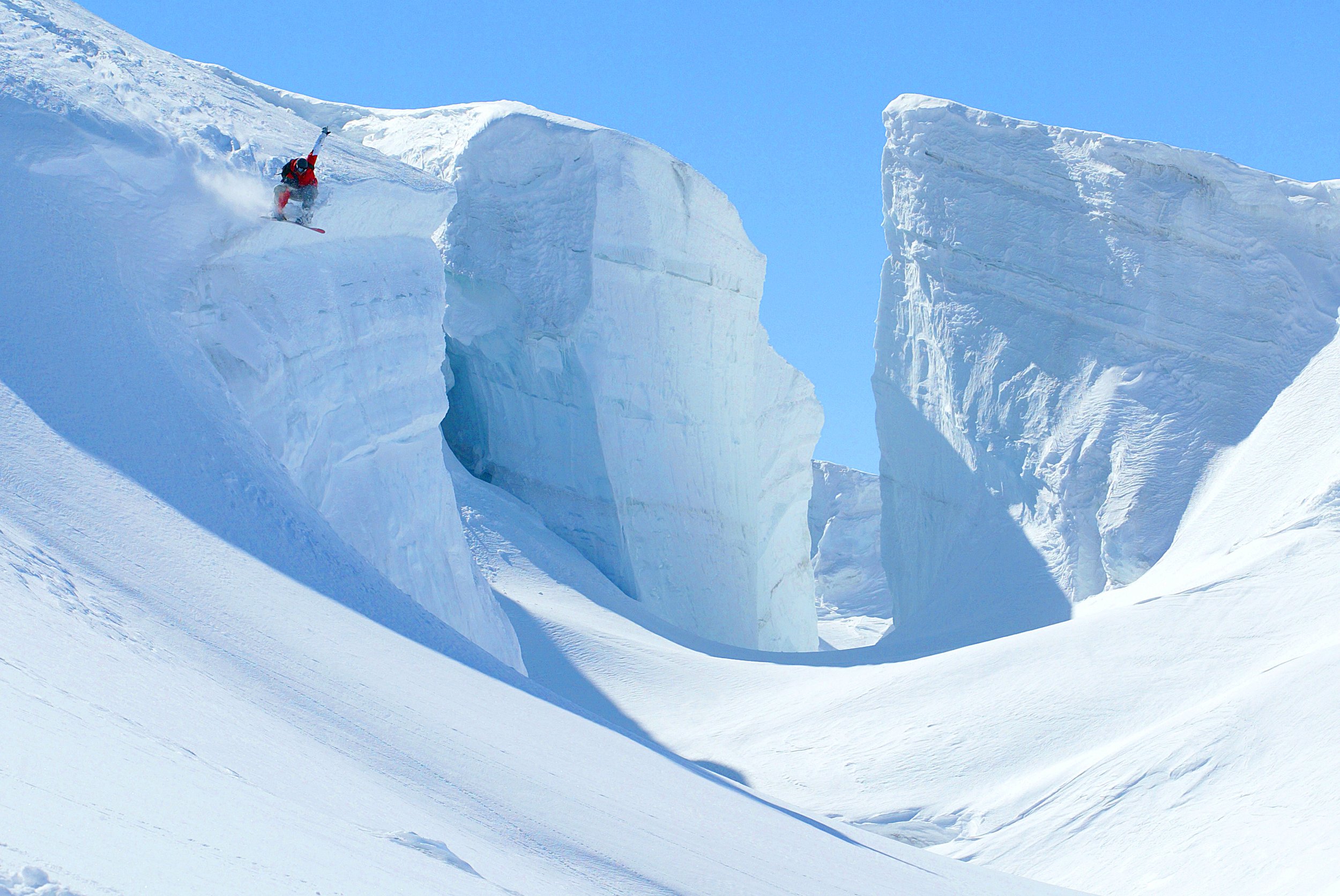 A person in red winter gear snowboarding on icy snow between large ice formations under a clear blue sky.