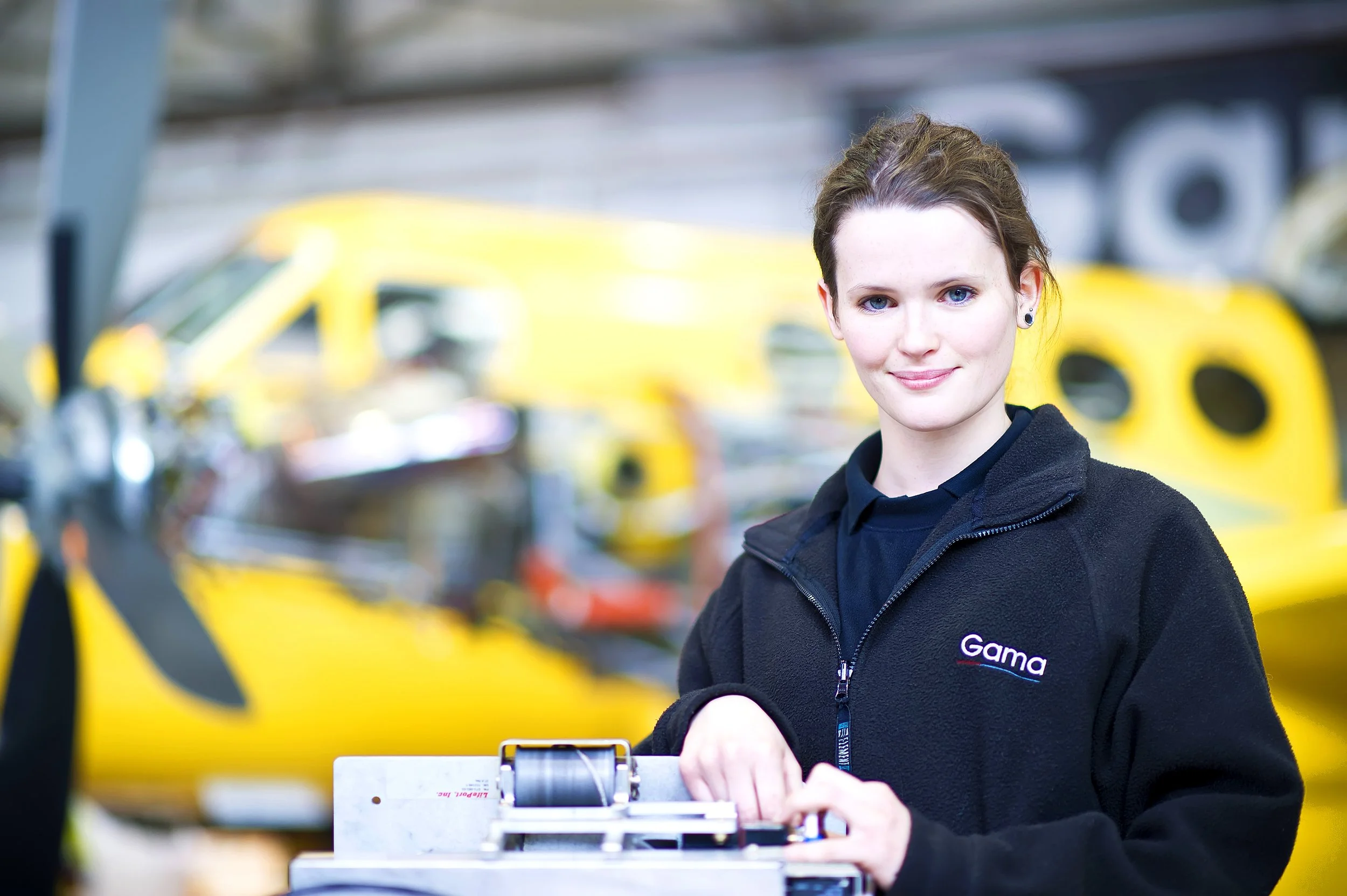 A young woman with short brown hair and blue eyes wearing a black jacket with a 'Gama' logo stands in front of yellow race cars at a racing event. She is smiling and appears to be operating a device or tool, with a blurred background of racing vehicl
