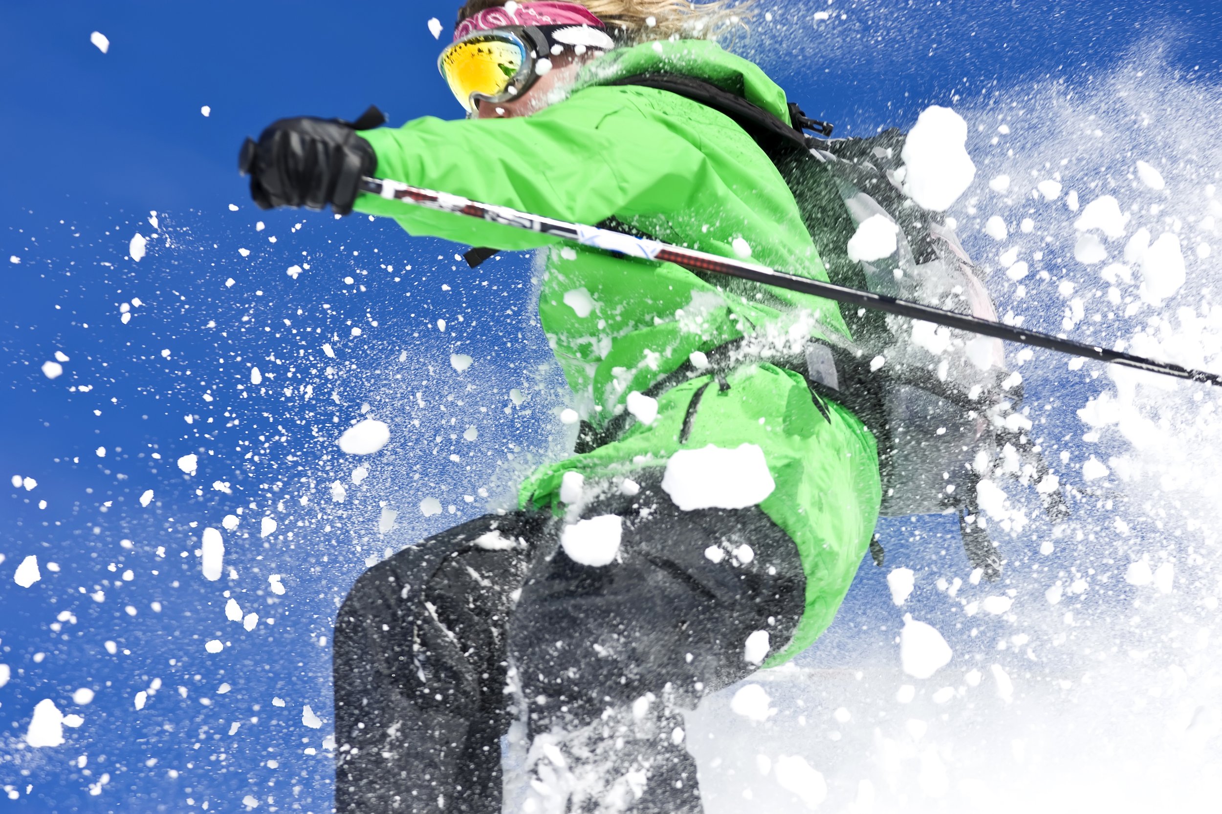 A person skiing on snow, wearing a green jacket, black gloves, orange-tinted goggles, and a pink headband, captured mid-movement with snow flying around.