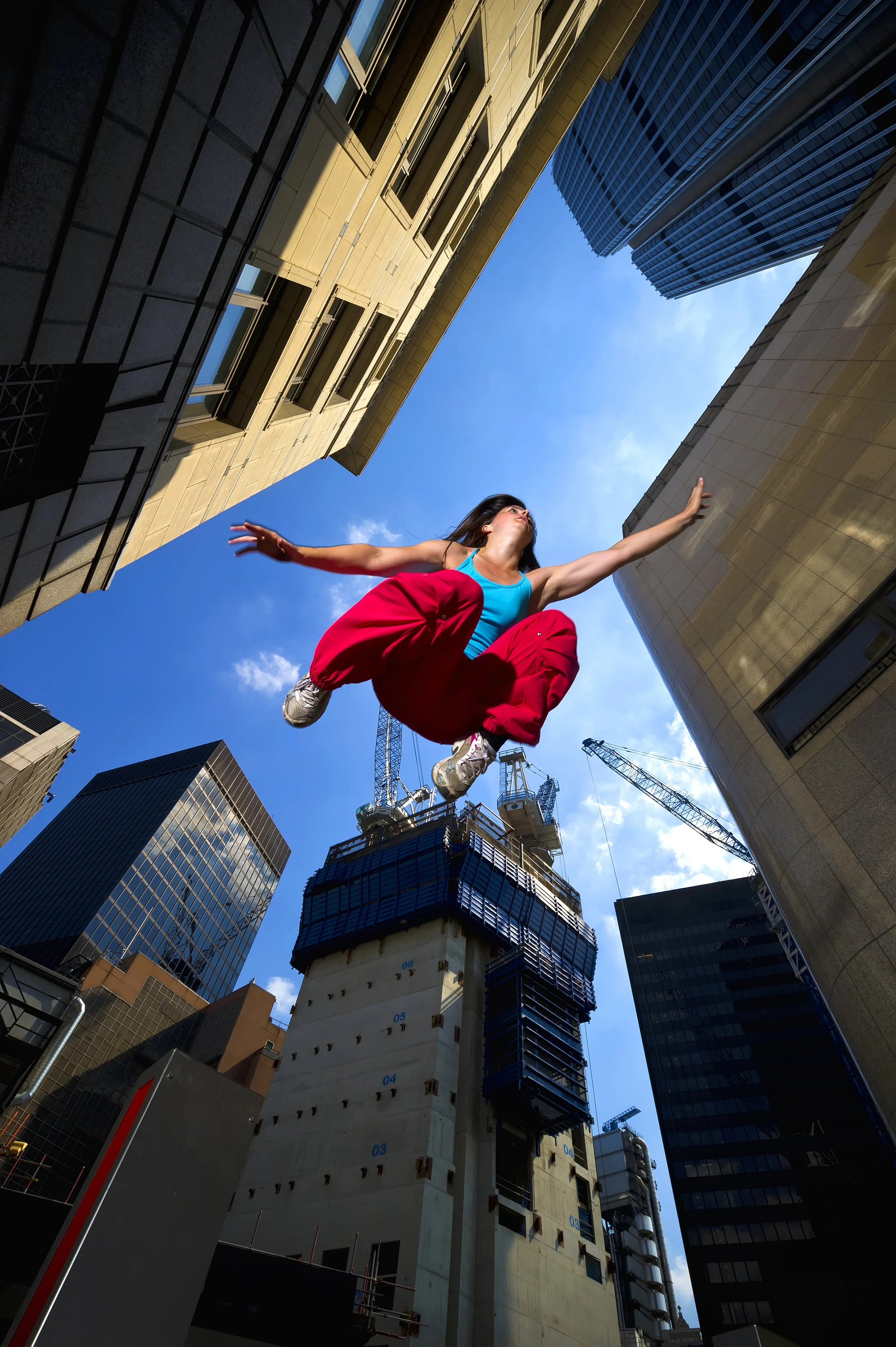 A woman dressed in a blue tank top and red pants is jumping in the air between tall skyscrapers in an urban cityscape, with construction cranes in the background and a clear blue sky above.