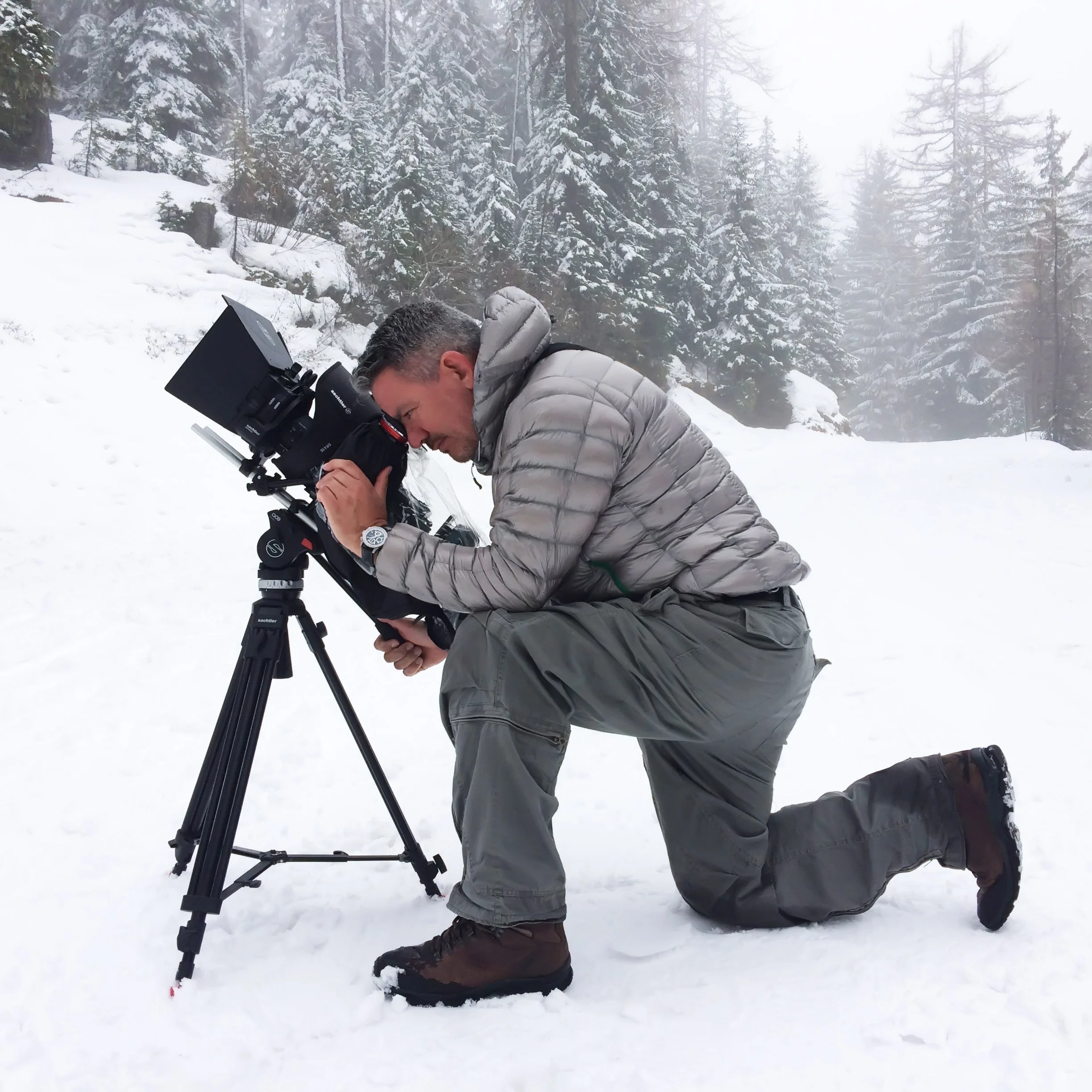 A man kneeling on snow while looking through a camera mounted on a tripod in a snowy forest.