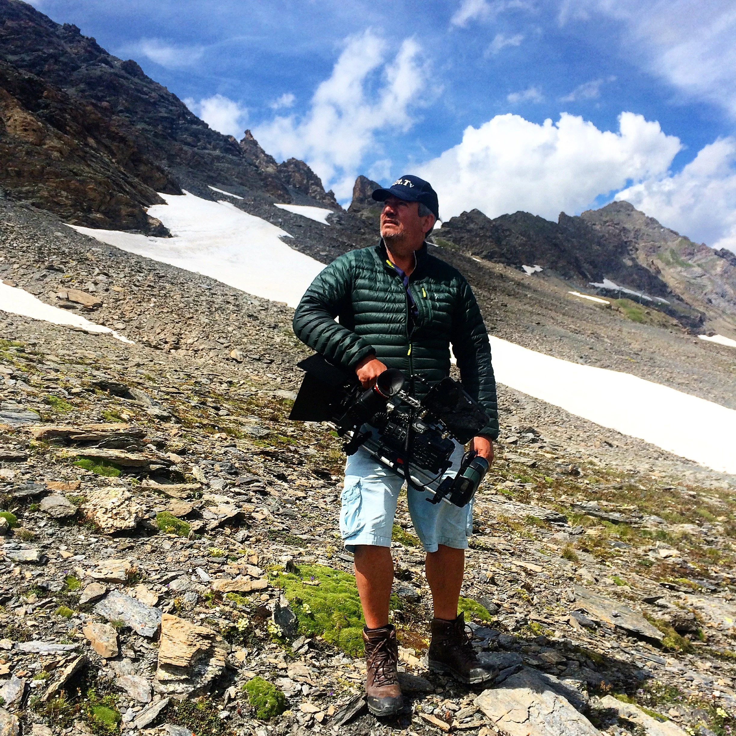 A man wearing a green jacket, white shorts, and hiking boots standing on rocky mountain terrain with patches of snow, under a partly cloudy blue sky.