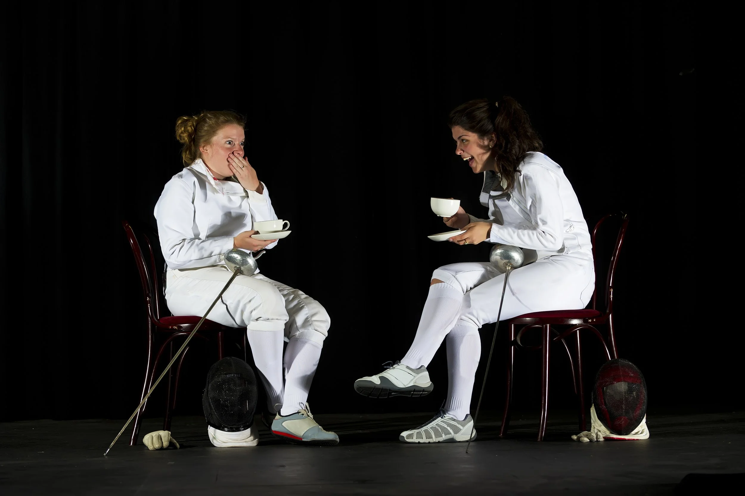 Two women in white outfits sitting on chairs against a black background, with cricket helmets, balls, and stumps at their feet. They are engaging in a humorous conversation, with one woman covering her mouth in surprise and the other smiling and hold