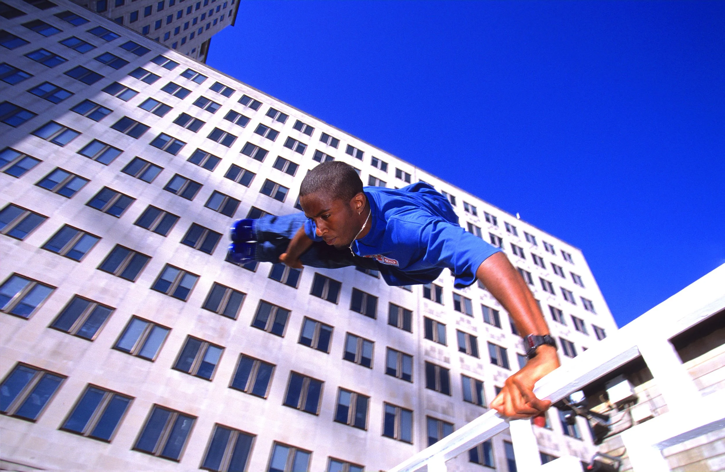 A man wearing a blue shirt and a necklace is leaning on a railing in front of a tall building with many windows, set against a bright blue sky, with a wide-angle perspective showing the skyscraper.