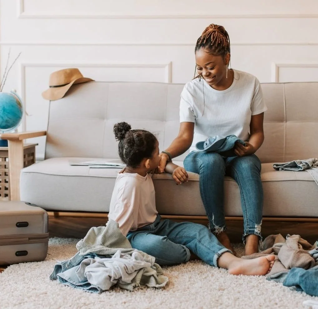 A woman and a young girl are playing together in a living room. The woman is sitting on a couch, smiling, while the girl is sitting on the floor, looking up at her. There is a pile of clothes and a suitcase on the floor, and a hat is resting on the back of the couch.
