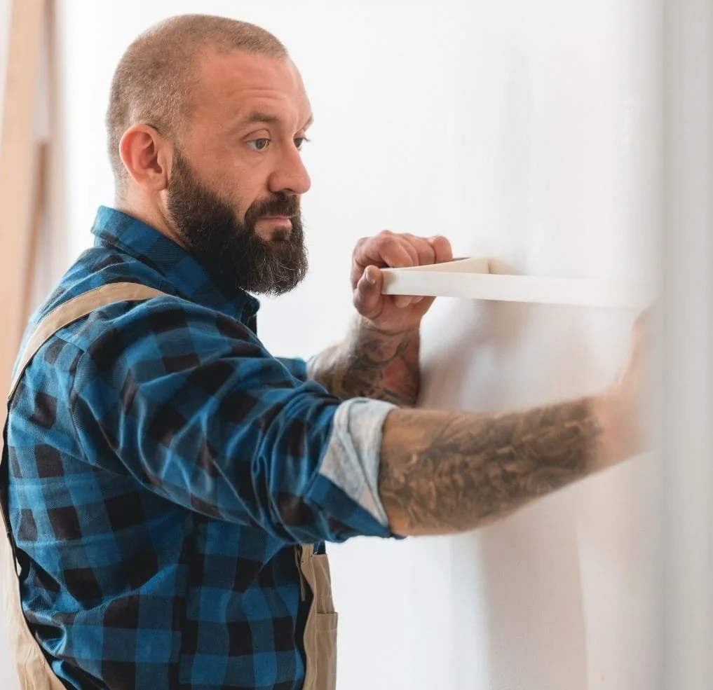 A tradesperson with a beard and tattoos installing a white shelf on a white wall.