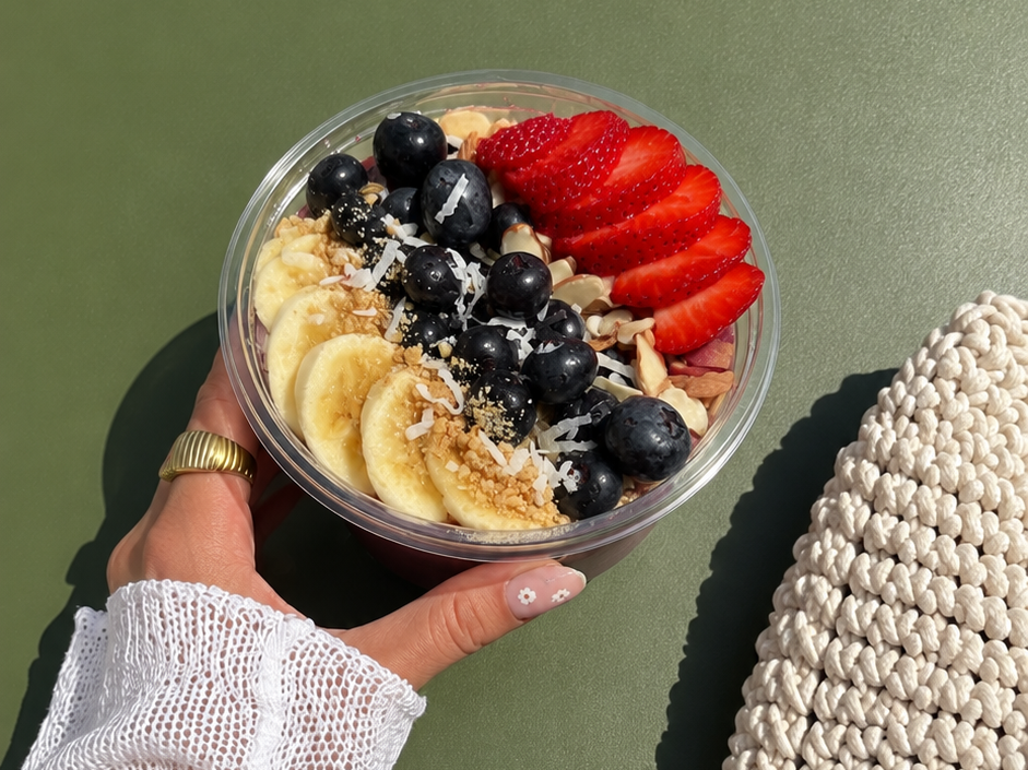 A clear plastic bowl filled with sliced bananas, blueberries, sliced strawberries, sliced almonds, shredded coconut, and crushed nuts, held by a person with a gold ring and a white lace sleeve.