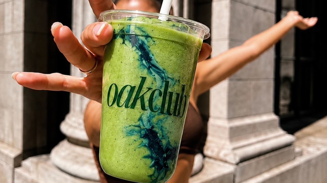 Person holding a green smoothie in a clear cup with a straw, outdoors on a sunny day.