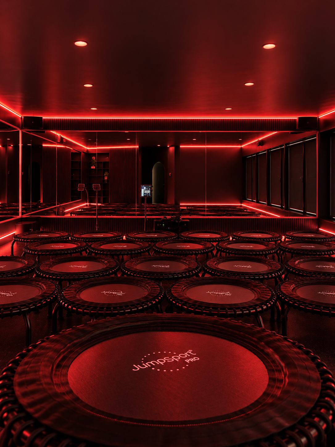 Indoor fitness studio with multiple mini trampolines arranged on the floor, illuminated by red LED lighting along the ceiling and walls, and a mirrored wall reflecting the room.