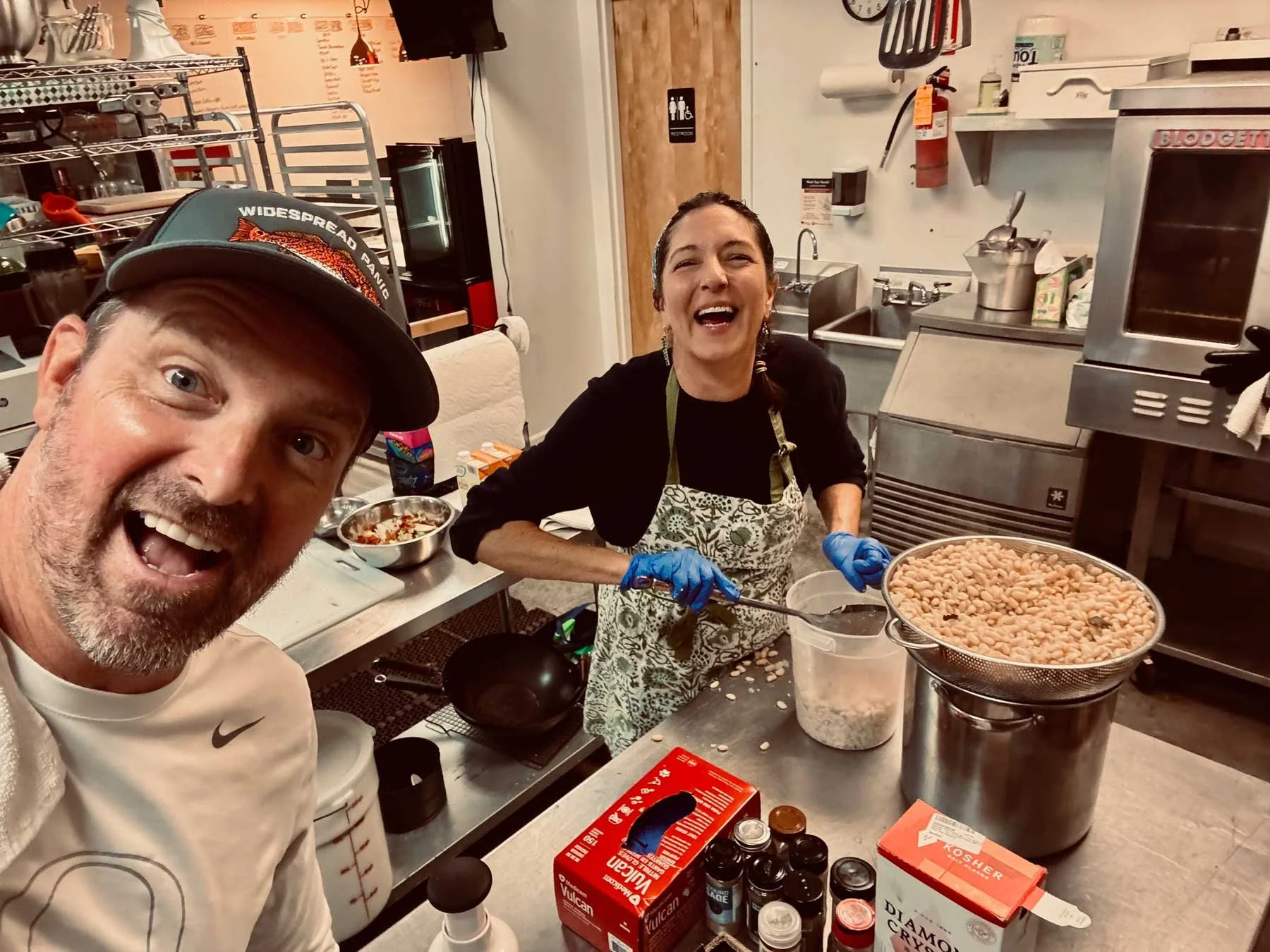 A man and a woman in a kitchen, smiling and working with food. The woman is scooping beans into a container, wearing blue gloves and an apron. The man is taking a selfie, wearing a hat and a white shirt. The kitchen has various utensils, boxes, and ingredients on the counters.