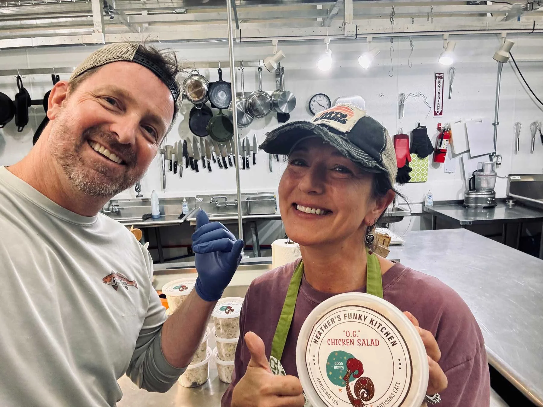 Two people smiling in a kitchen or food preparation area. The person on the left is a man wearing a beige cap backwards and a white t-shirt, while the person on the right is a woman wearing a baseball cap, earrings, an apron, and holding a container of chicken salad. The background shows hanging pots, pans, knives, and kitchen supplies.